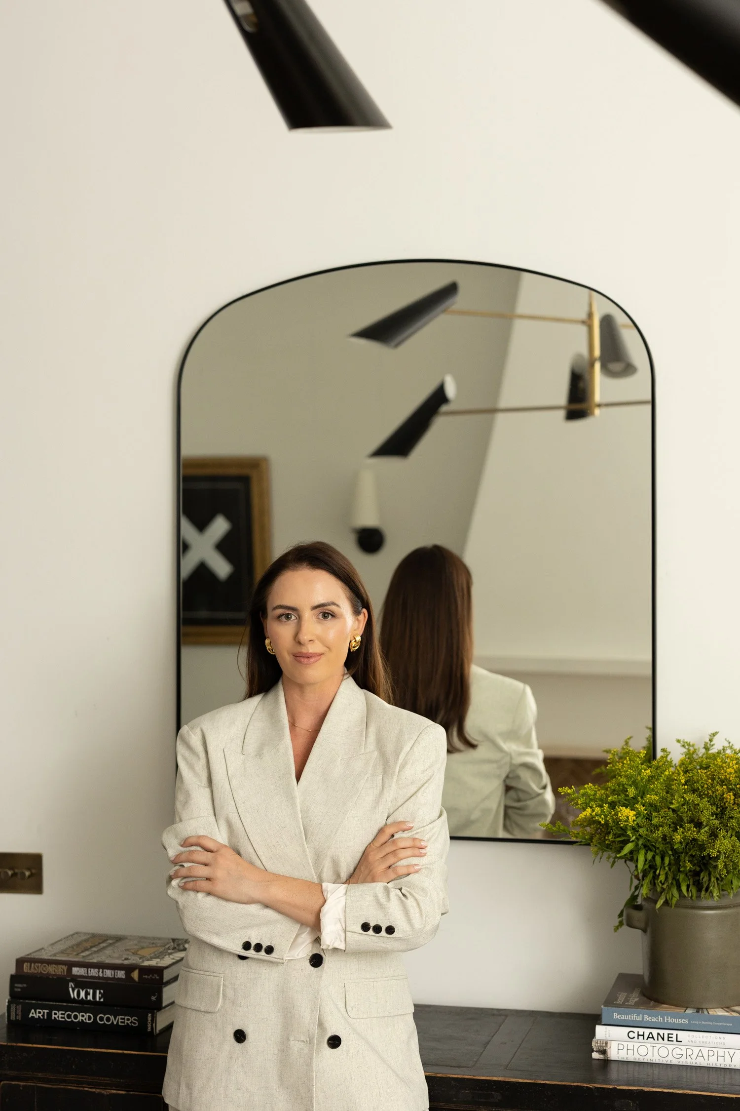 A woman with brown hair, dressed in a cream-colored blazer, standing with crossed arms in front of a mirror in a modern interior setting, with a desk, books, and a potted plant visible.