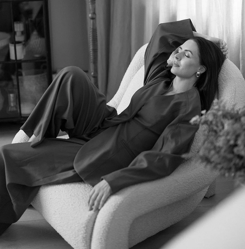 A woman relaxing on a sofa with her eyes closed and a serene expression, wearing a long-sleeved blouse and pants, in a cozy indoor setting.
