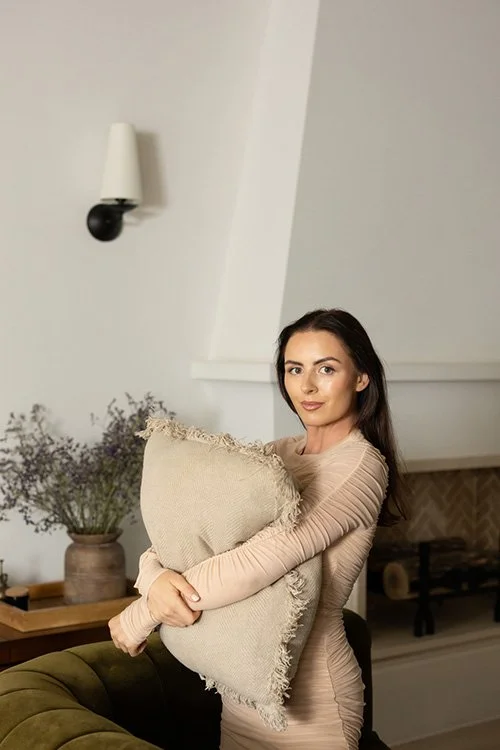 A woman with long dark hair holding a beige pillow in a living room.