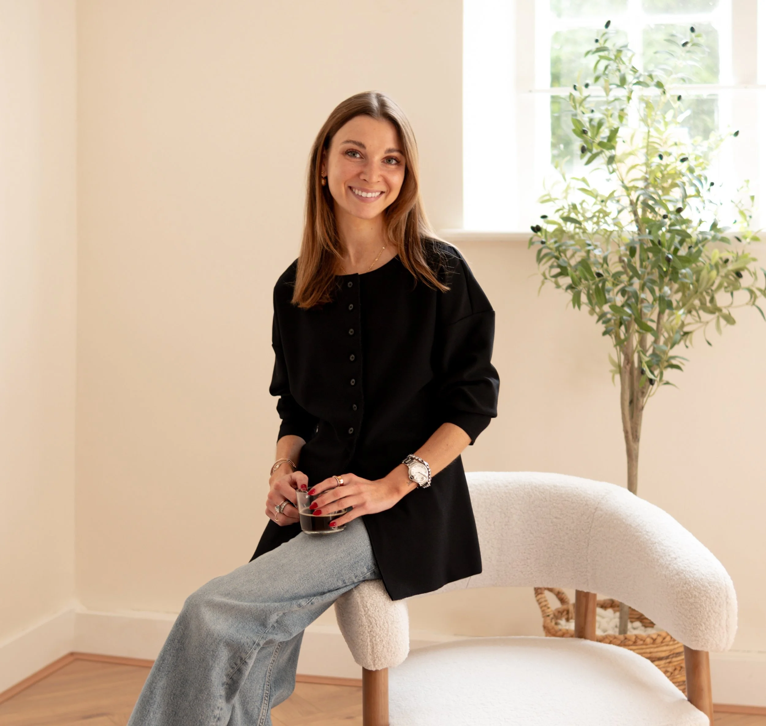 A woman in a white jacket and black skirt posing indoors, with clothing on hangers and a large potted plant in the background.