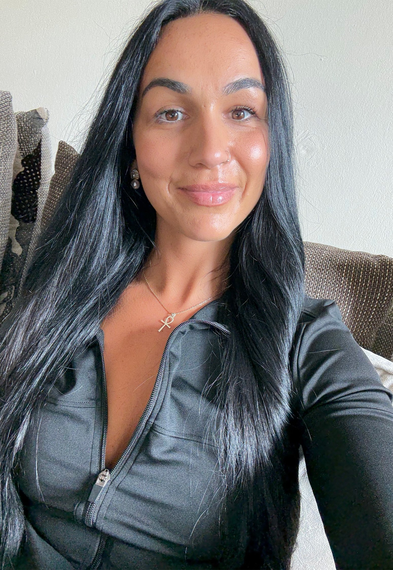 A woman with long black hair, wearing a black zip-up jacket, a necklace with an ankh pendant, and pearl earrings, smiling while sitting on a brown couch with textured cushions in the background.