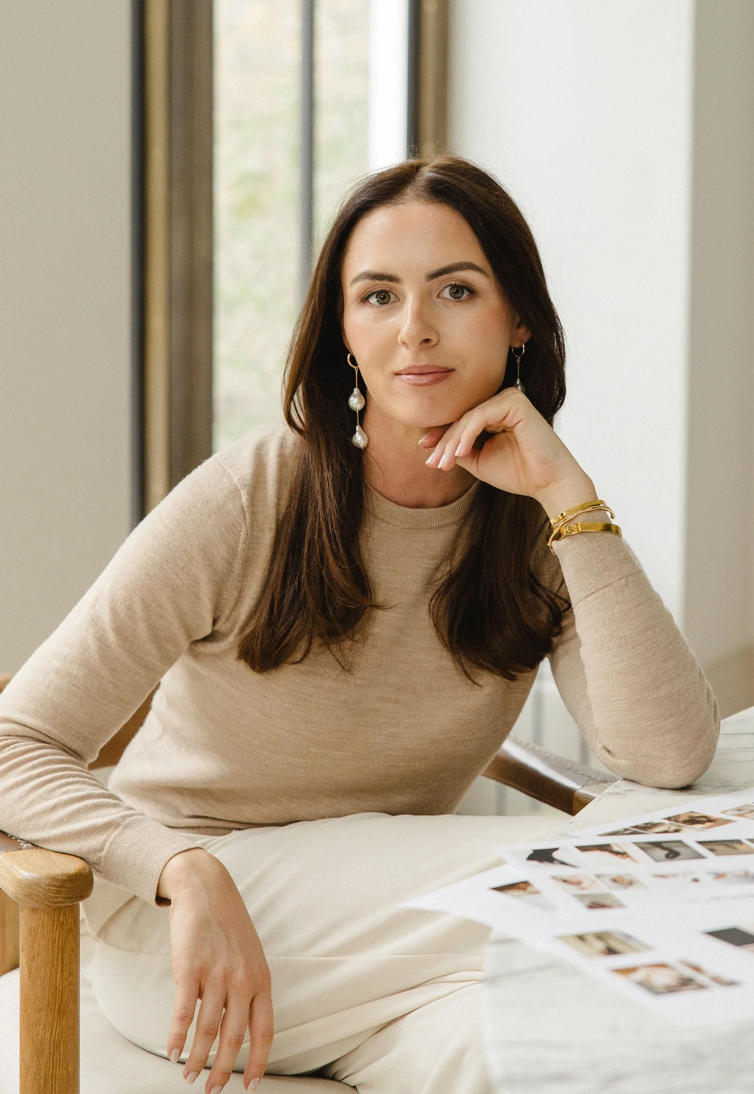A woman with long dark hair and earrings sitting at a table, resting her chin on her hand, with photographs spread out in front of her.