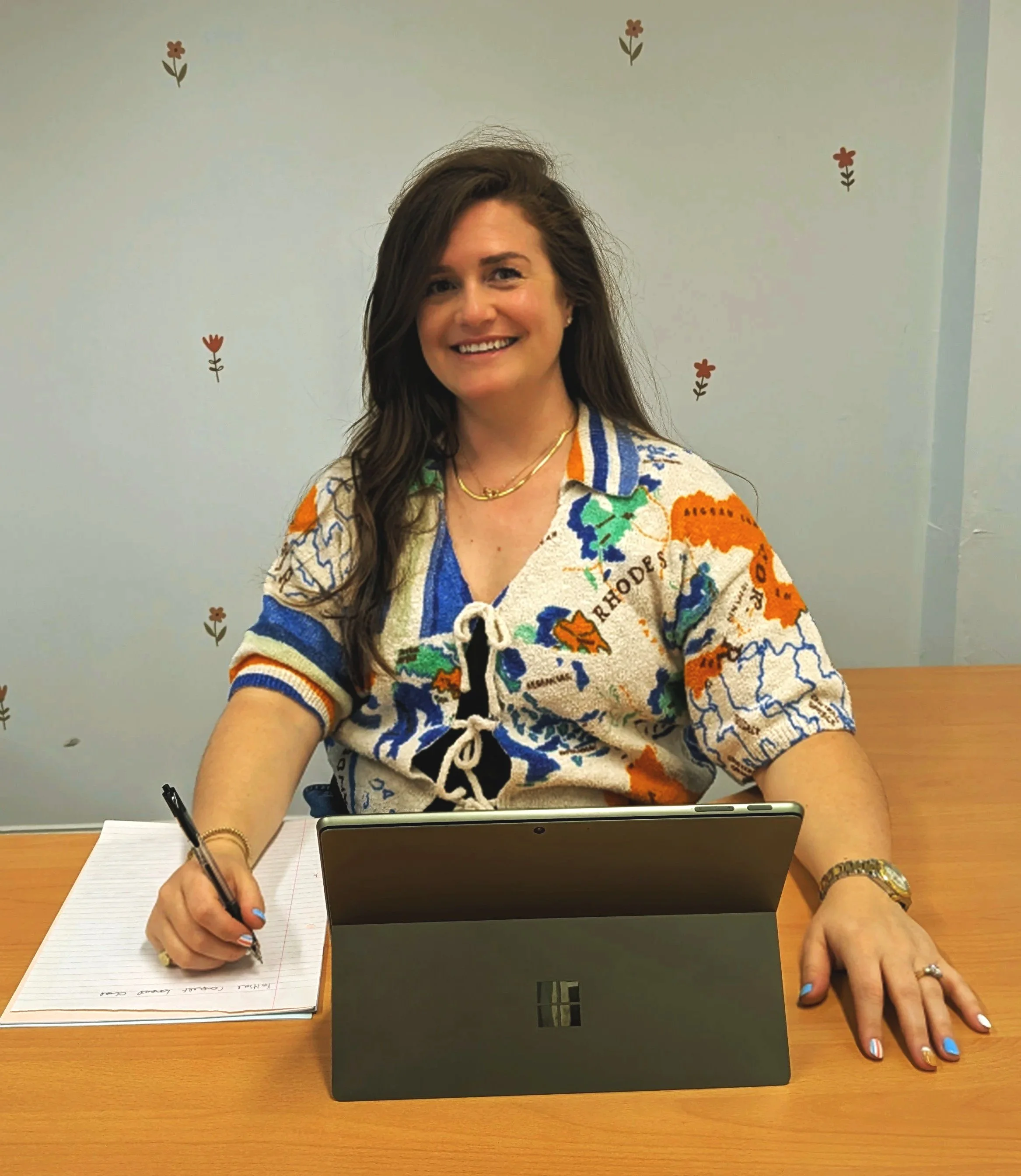 Rachel Maunsell is sitting at a wooden table in front of a laptop. She is writing in a notebook with a pen. The background features a plain wall with small flower decorations.
