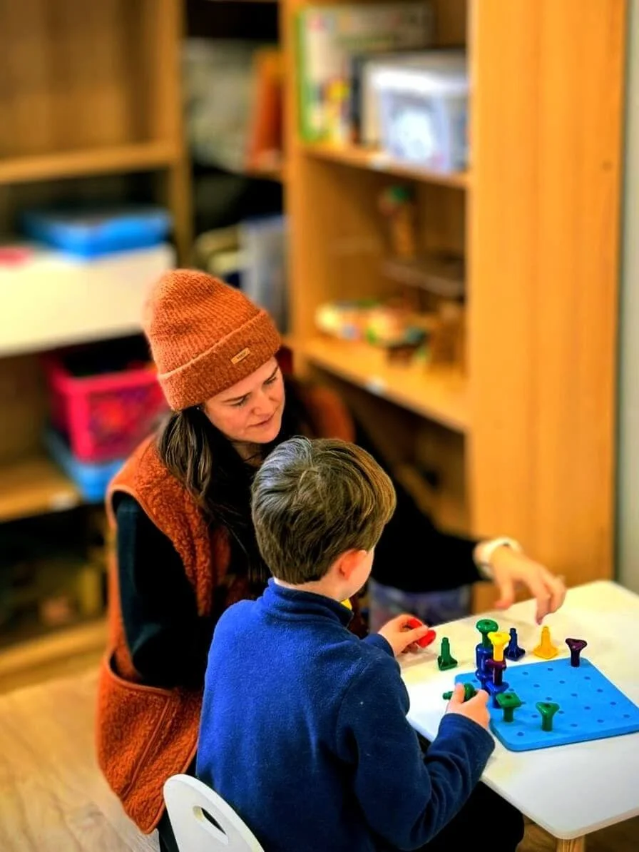 A woman and a young boy playing a board game with colorful tokens at a small table in a room with wooden bookshelves.