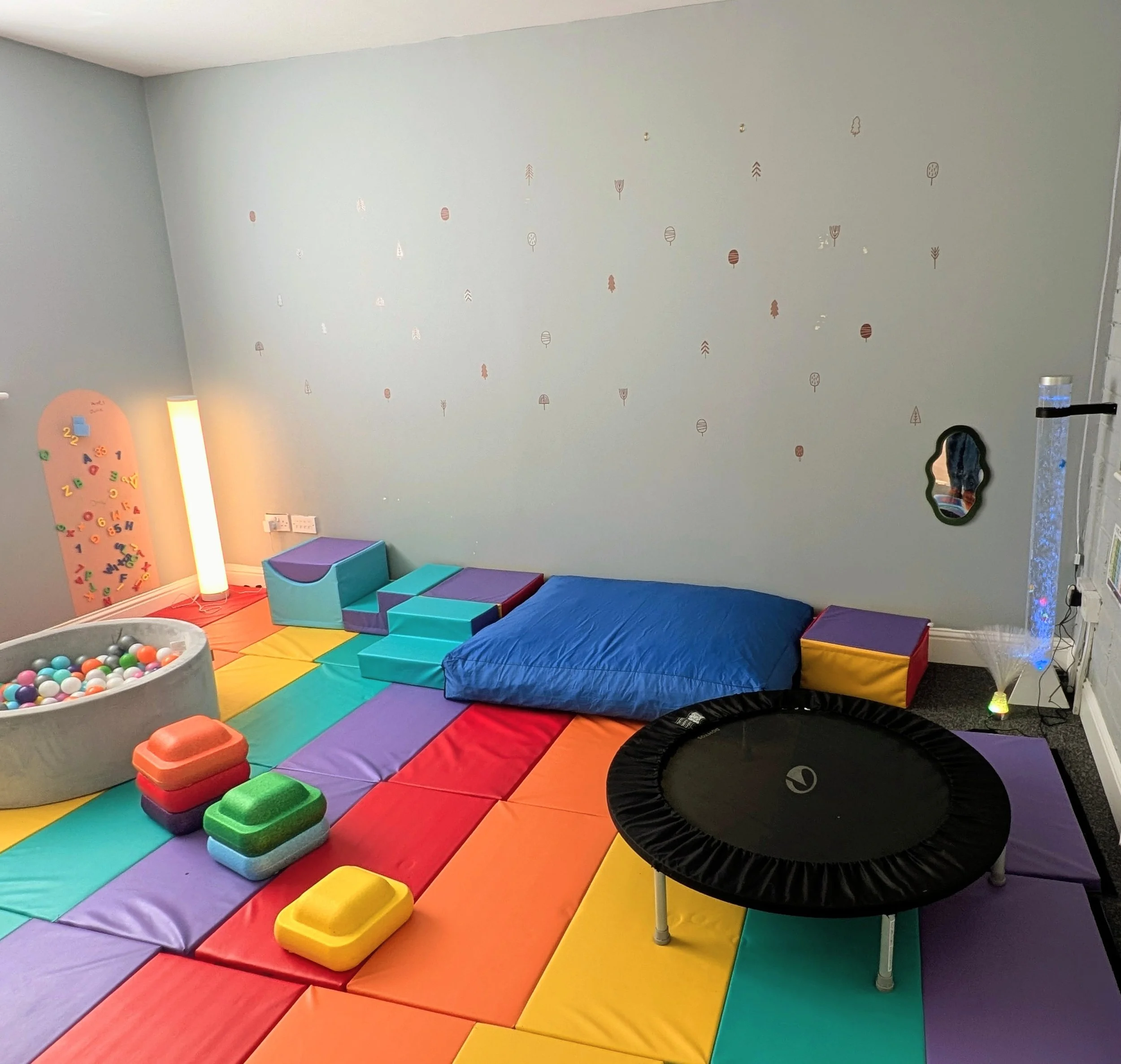 Colorful indoor playroom with a rainbow foam mat, a mini trampoline, a small trampoline, foam blocks, a ball pit, and wall decorations, designed for children.