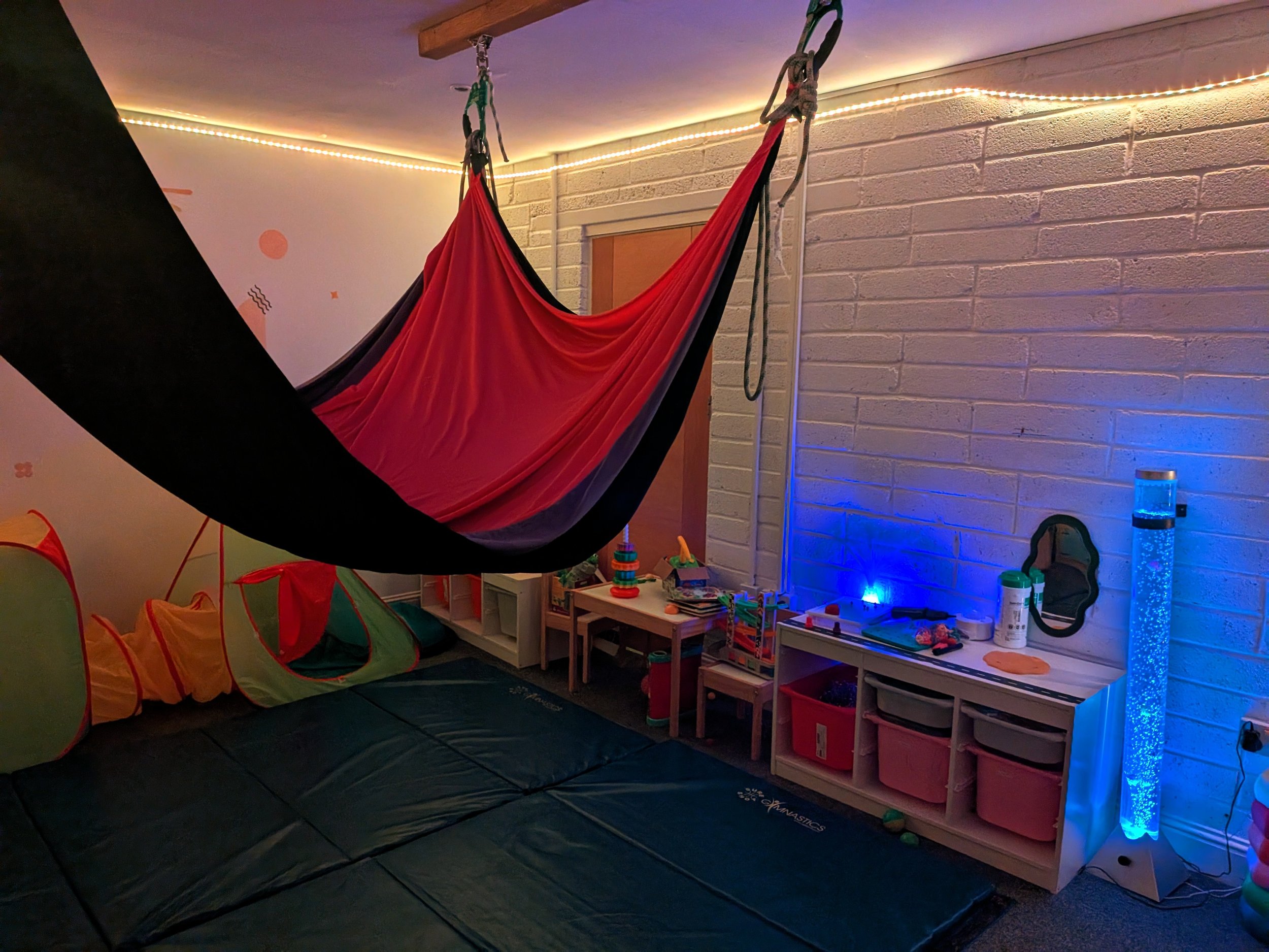 Indoor playroom with a black and red hammock, a tent, foam mats, and shelves with toys. Wall has white bricks and colorful lighting.
