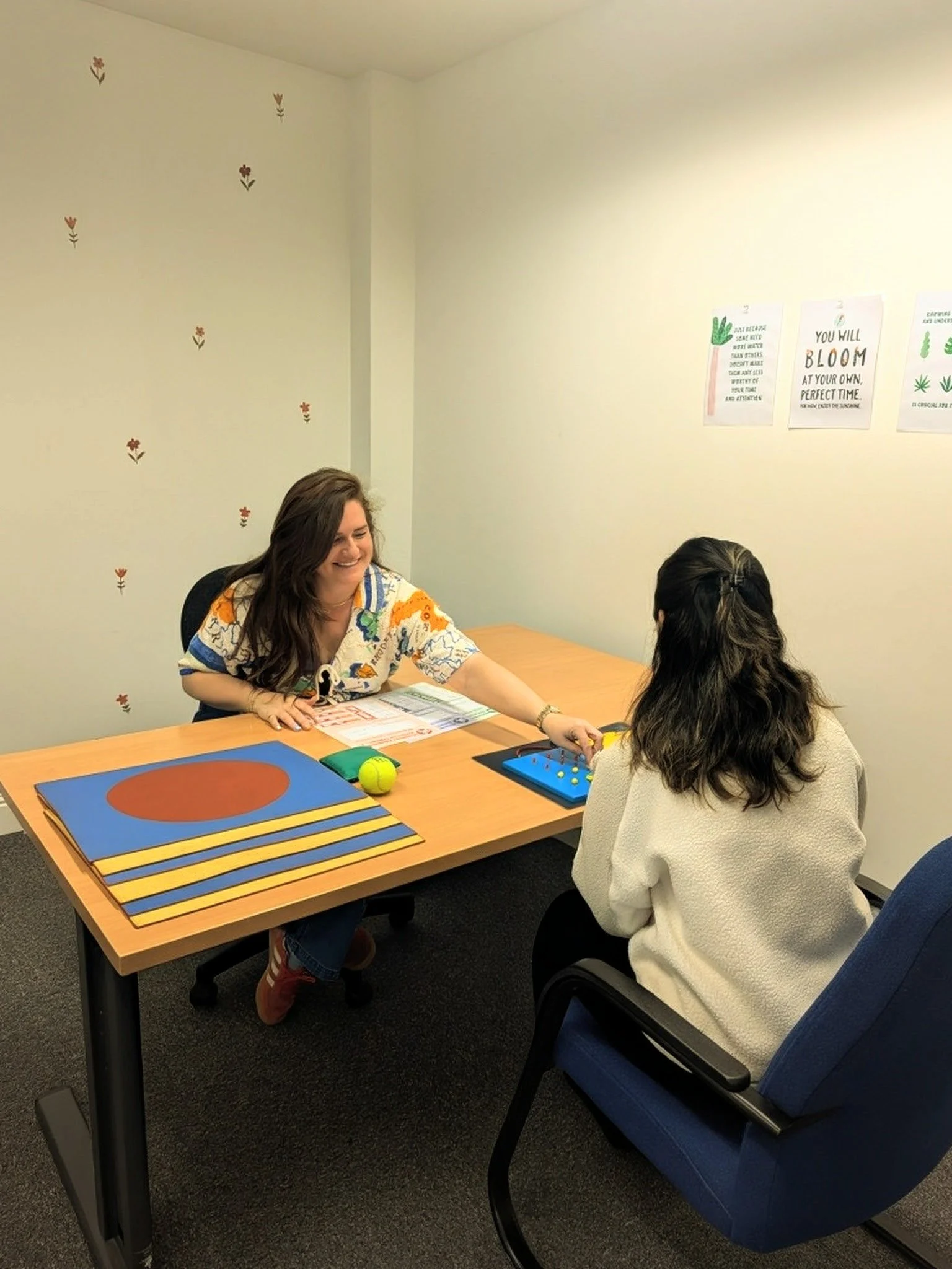Two women sitting at a conference table in an office, engaged in conversation. One woman is smiling and reaching over the table. The table has documents, a tennis ball, and framed photos or designs. The wall behind them is decorated with small flower decals and motivational posters.