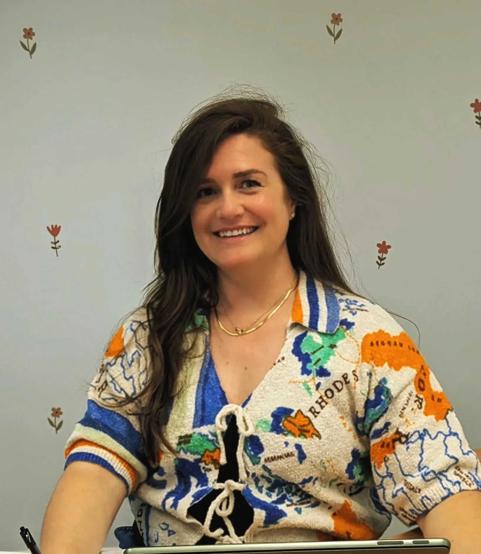 A woman with long dark hair smiling, wearing a colorful sweater with a world map design, sitting at a table with a pen and tablet in front of her, against a light-colored wall with small floral decorations.