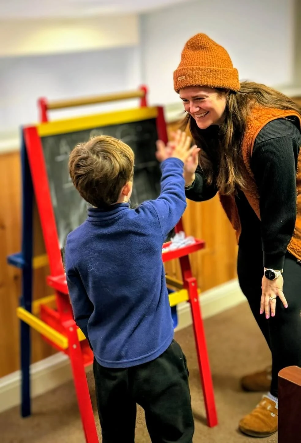 A woman wearing a brown beanie and orange vest high-fives a young boy in a blue sweater in a classroom.