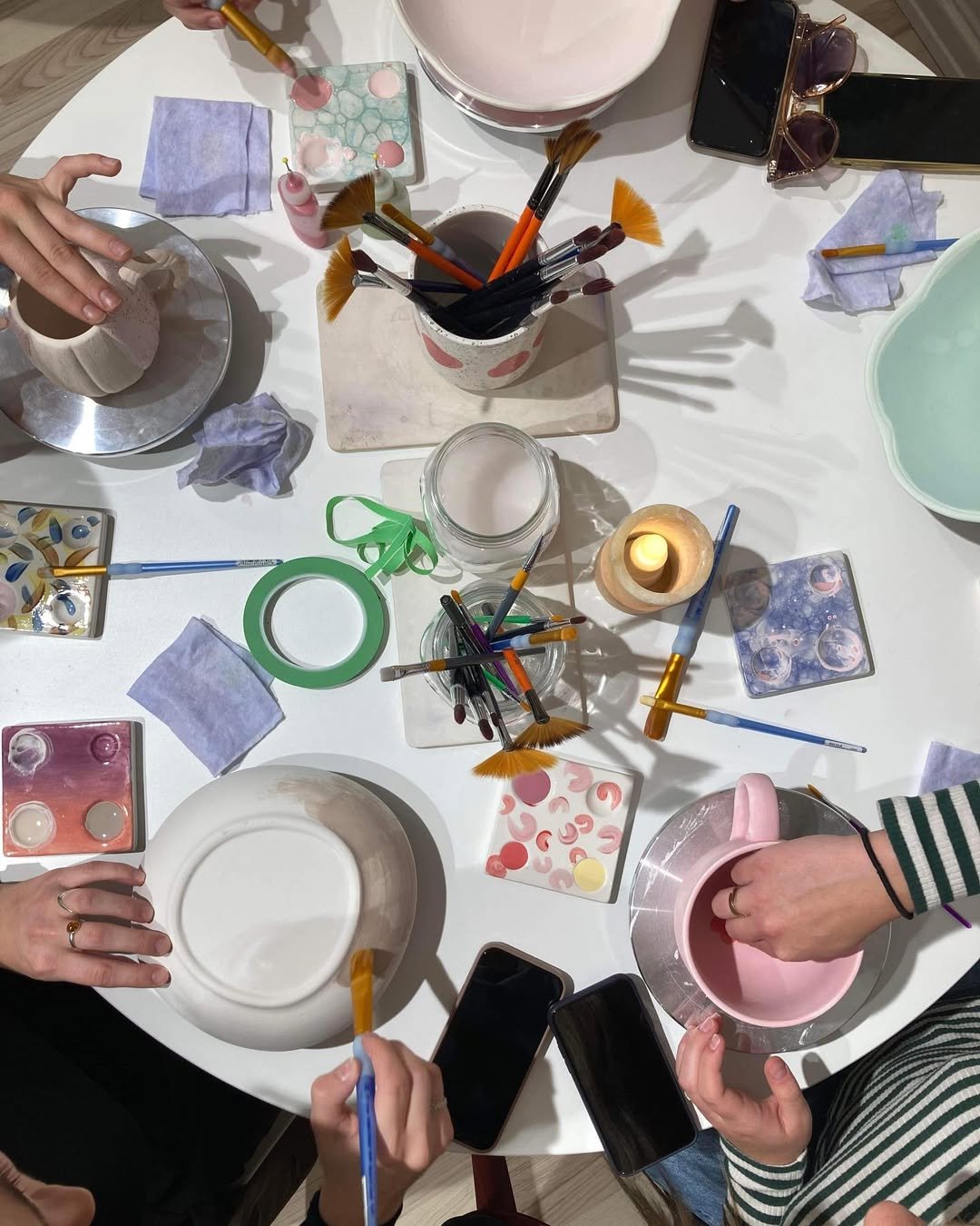 Billede af en gruppe mennesker, der laver håndværk eller dekorationer på et hvidt bord med forskellige farvede maling, børster, plastikskabeloner, lys, papir og telefoner.