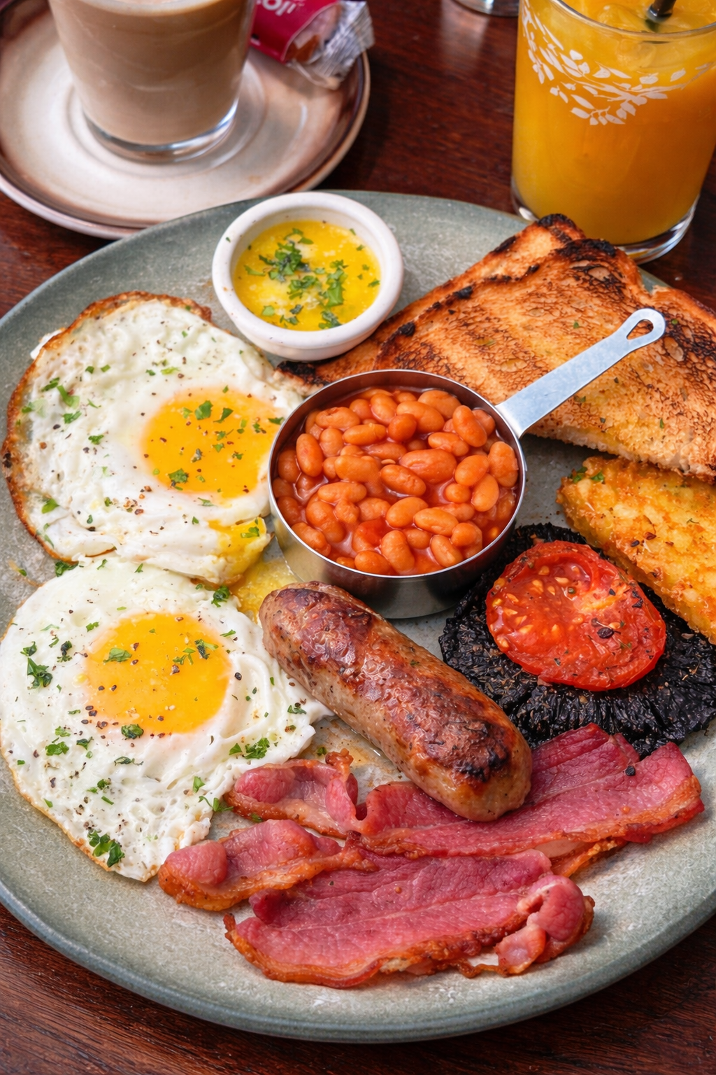 A breakfast plate with fried eggs, baked beans, sausage, bacon, grilled tomato, hash browns, and toast. A small dish of melted butter with herbs is also on the plate. Drinks include a glass of orange juice, a cup of coffee on a saucer, and a tall glass of orange drink.