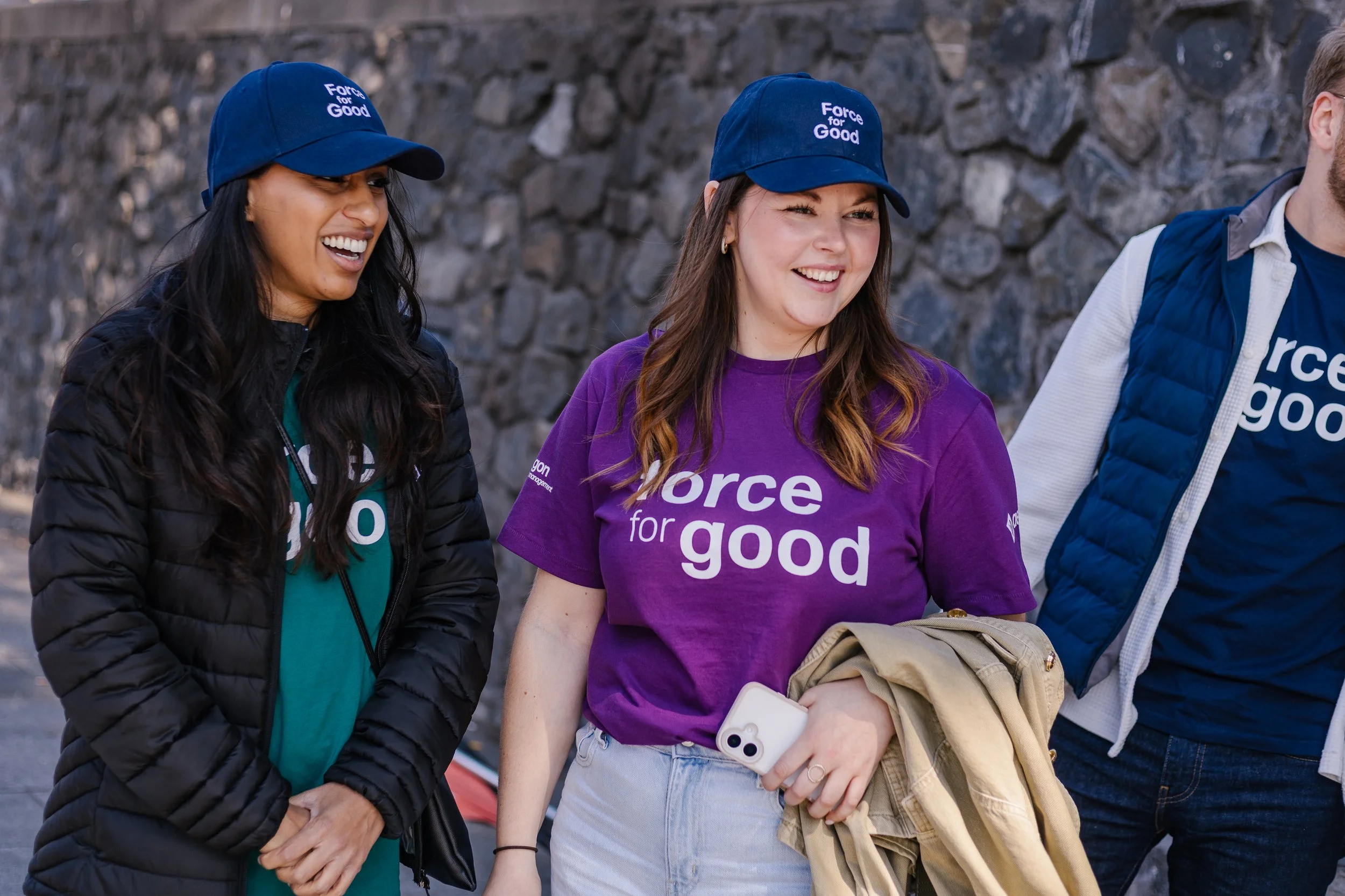 Three smiling people wearing 'Force for Good' hats and shirts, standing outdoors near a stone wall.