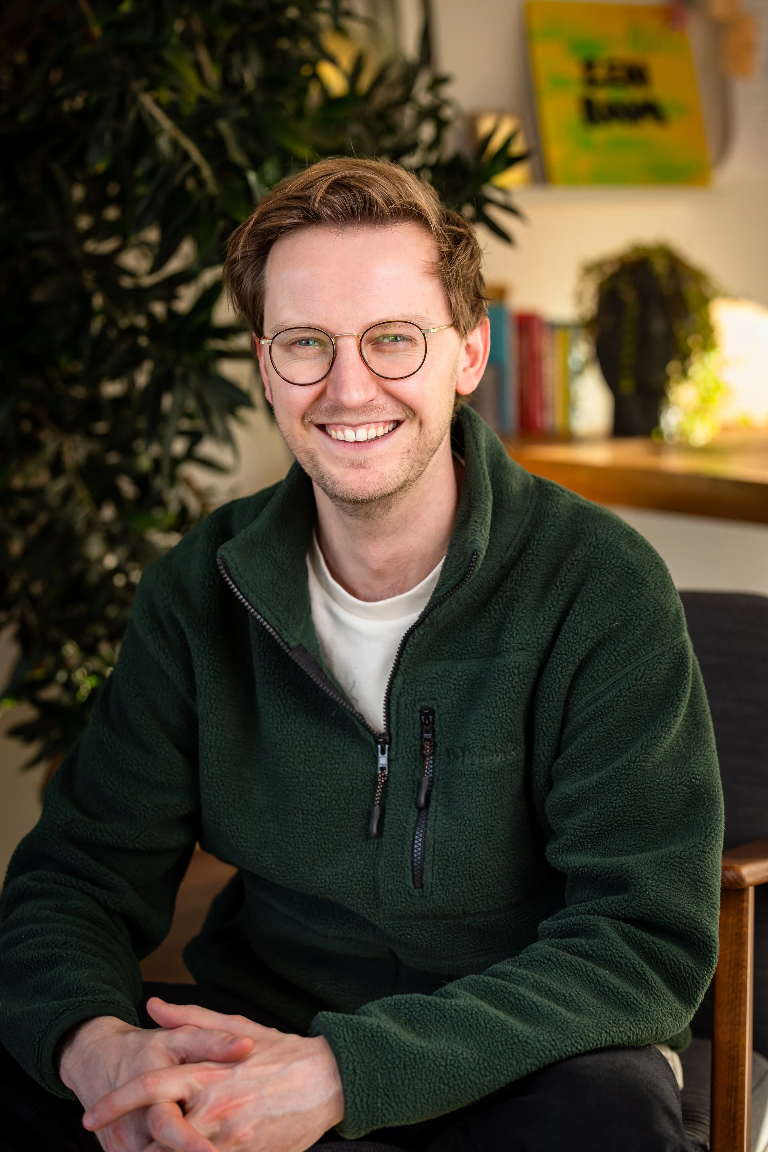 A smiling man with glasses and brown hair in a green fleece jacket, sitting indoors with a plant, bookshelf, and colorful artwork in the background.