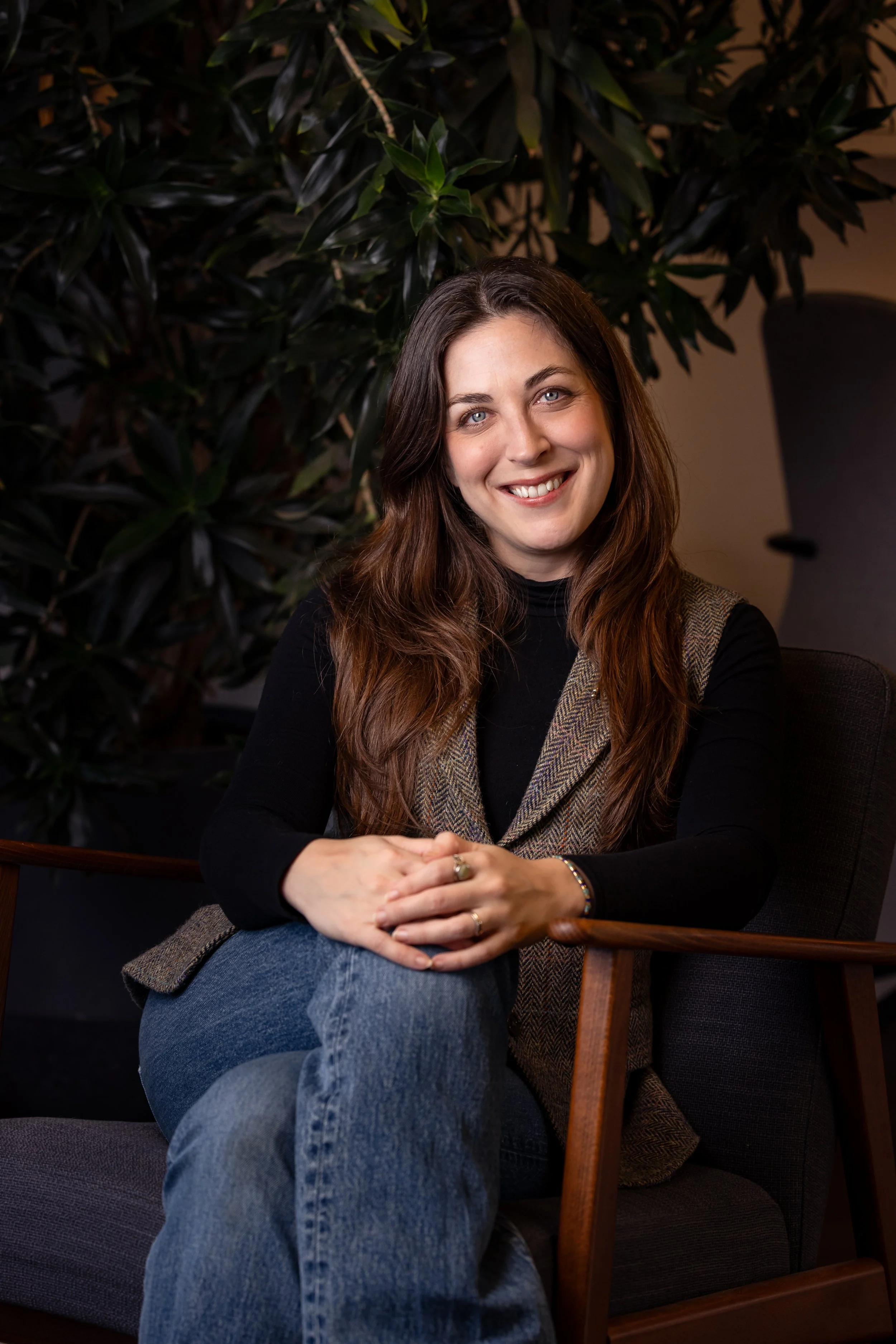A woman with long brown hair and blue eyes sitting on a wooden chair with black cushions, smiling at the camera in front of a large leafy green plant.