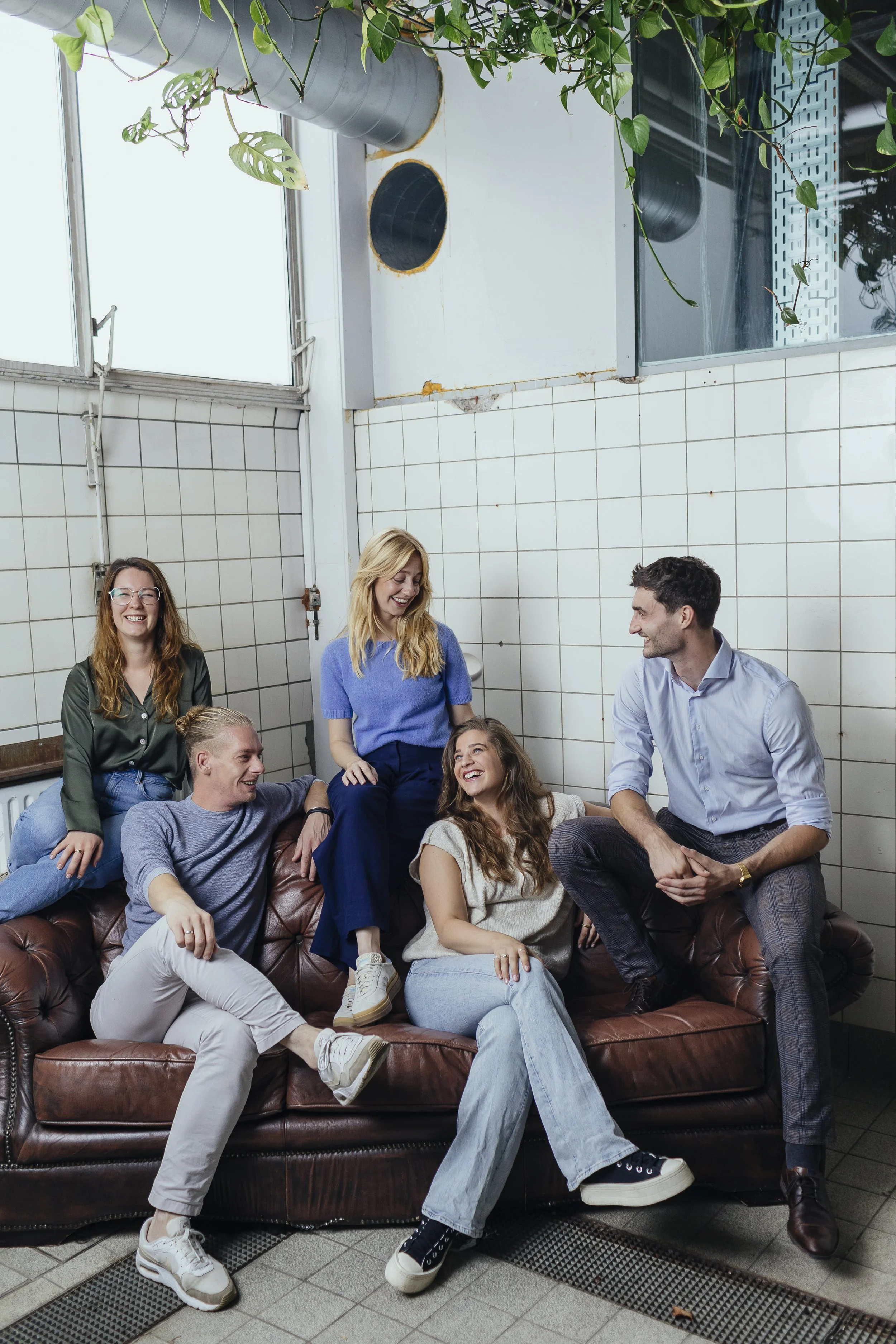 Group of five people laughing and chatting while sitting on and around a brown leather sofa in a partly industrial, tiled room with large windows and greenery hanging from above.