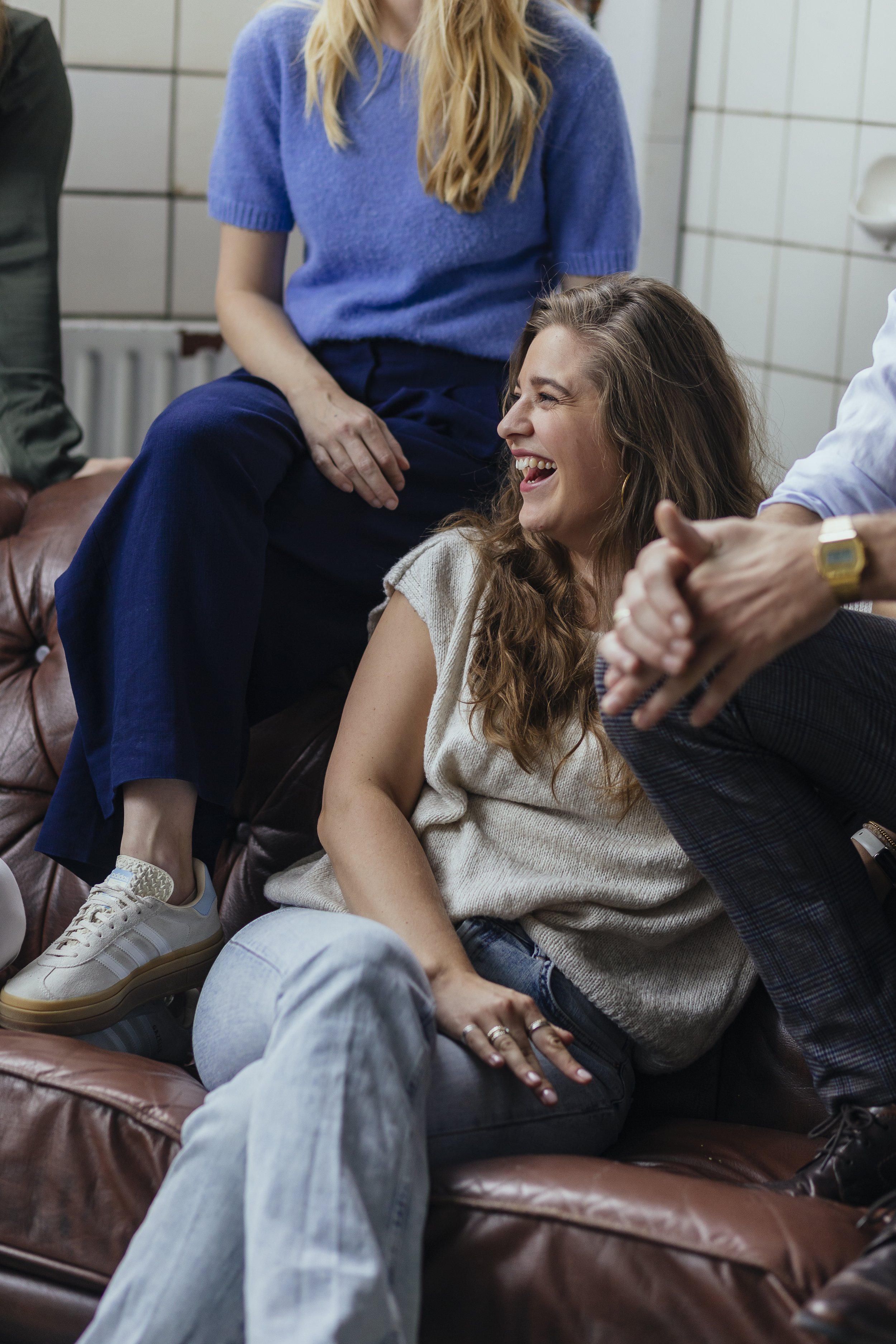 A group of people laughing and having fun together indoors, sitting on a brown leather couch and a tiled wall in the background.