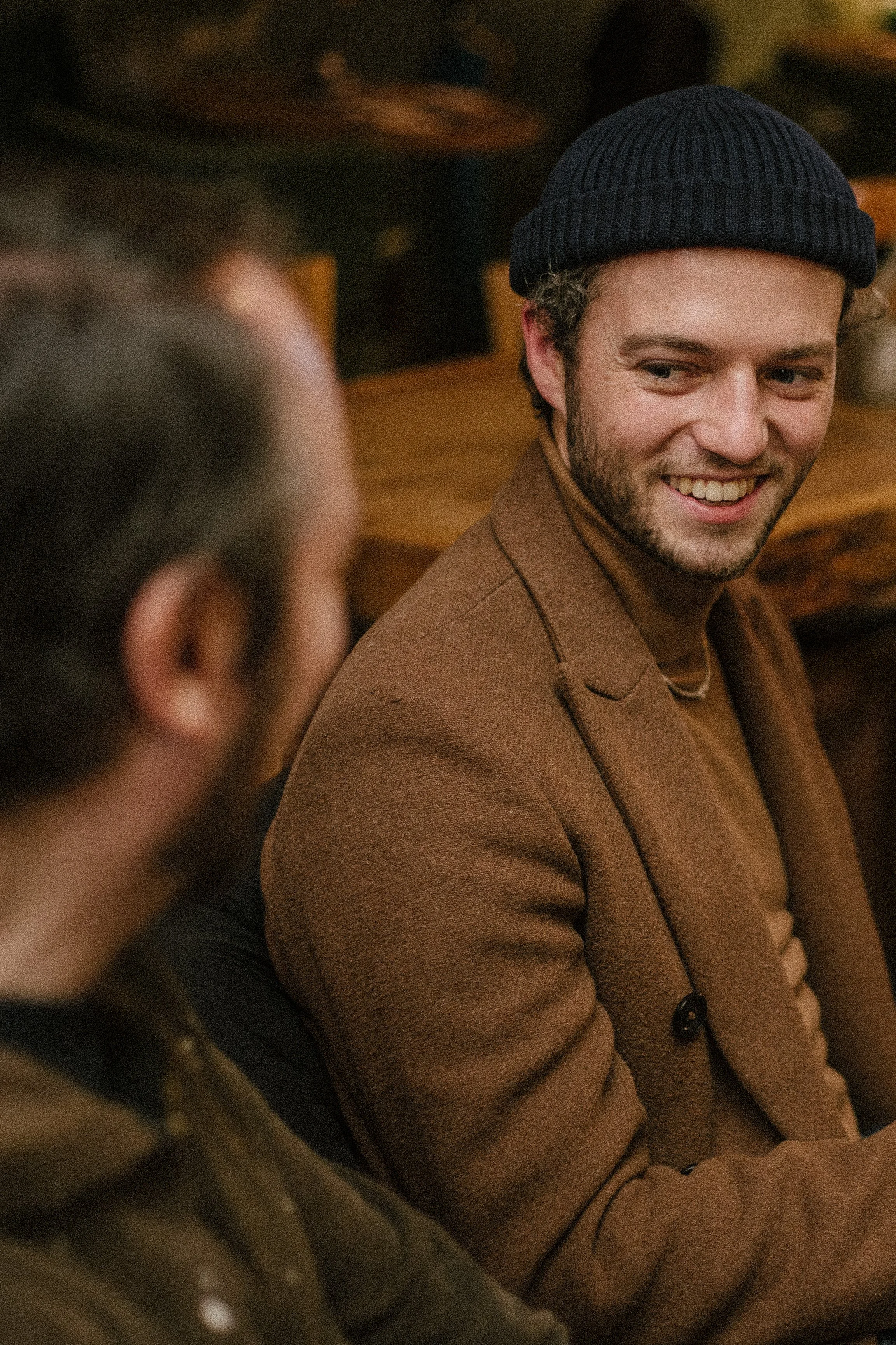 Two men sitting at a table in a cozy, wood-paneled setting, engaged in conversation. One man is smiling, wearing a brown blazer and a navy beanie, while the other, with blurred features in the foreground, has dark hair and a beard.