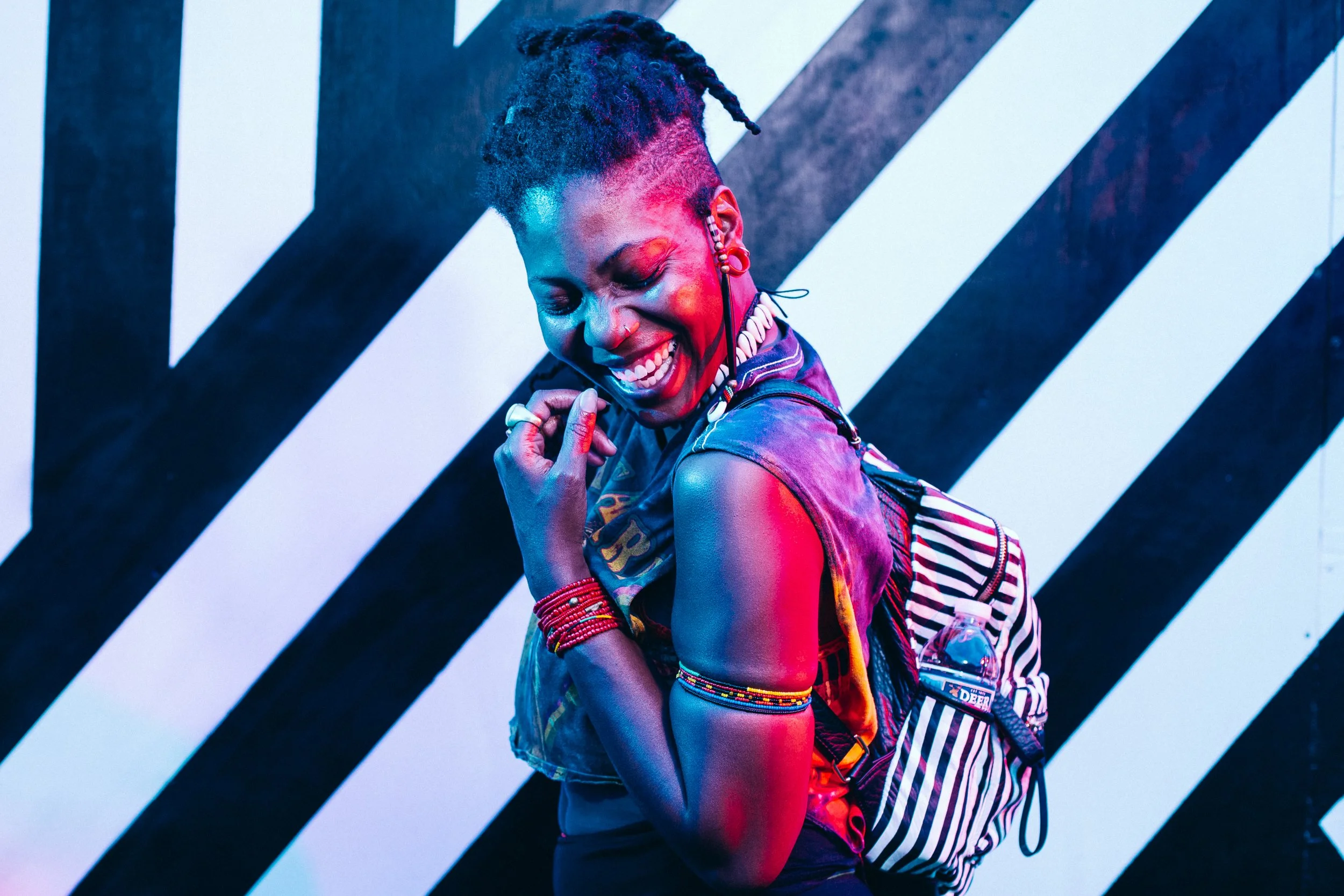 A woman with short, asymmetrical hair styled in dreadlocks, smiling and looking down, wearing colorful jewelry and a striped backpack, against a black-and-white diagonal striped background with pink and blue lighting.