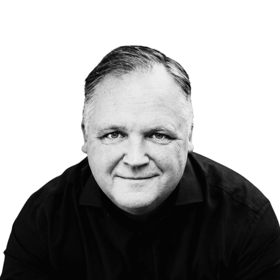 Black and white portrait of a middle-aged man with short, slicked-back hair, wearing a dark shirt, smiling slightly, against a black background.