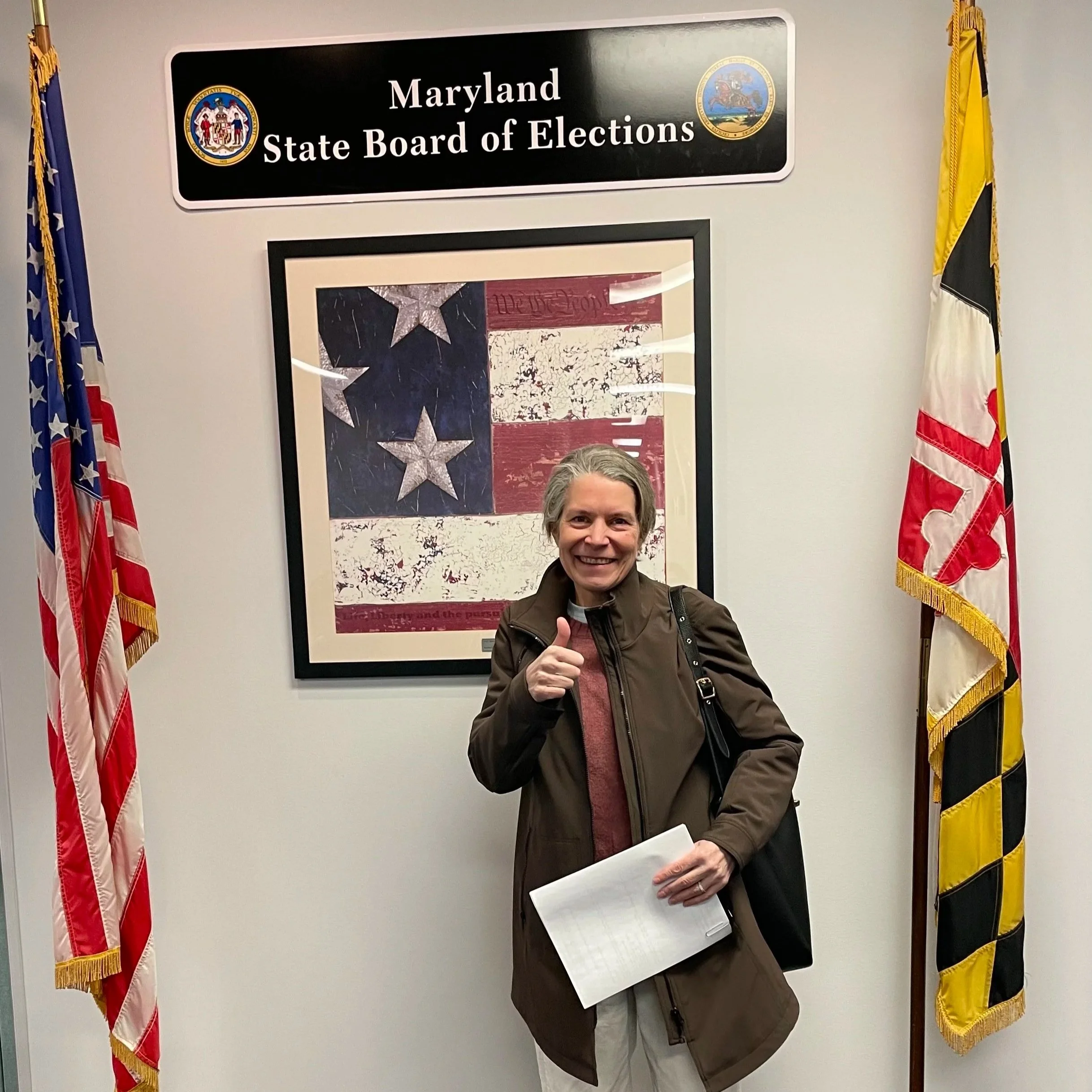 Kate Stein smiling and giving a thumbs-up pose in front of a Maryland State Board of Elections sign, flanked by American flags.