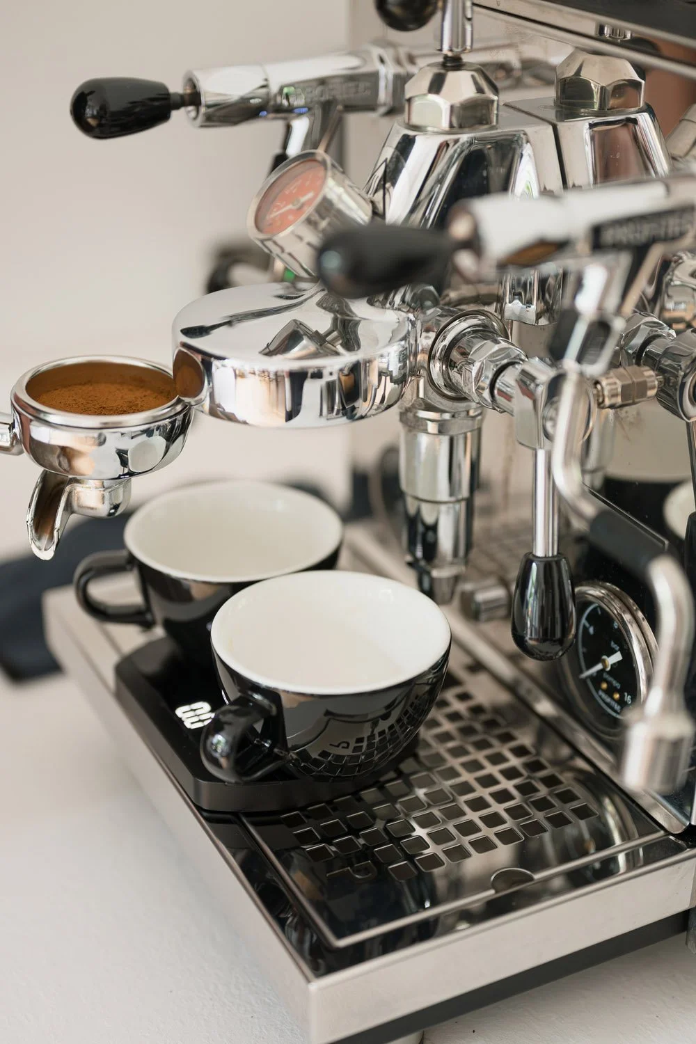 A shiny espresso machine with black handles brewing coffee. There are two black and white cups beneath the spouts, one filled with ground coffee.