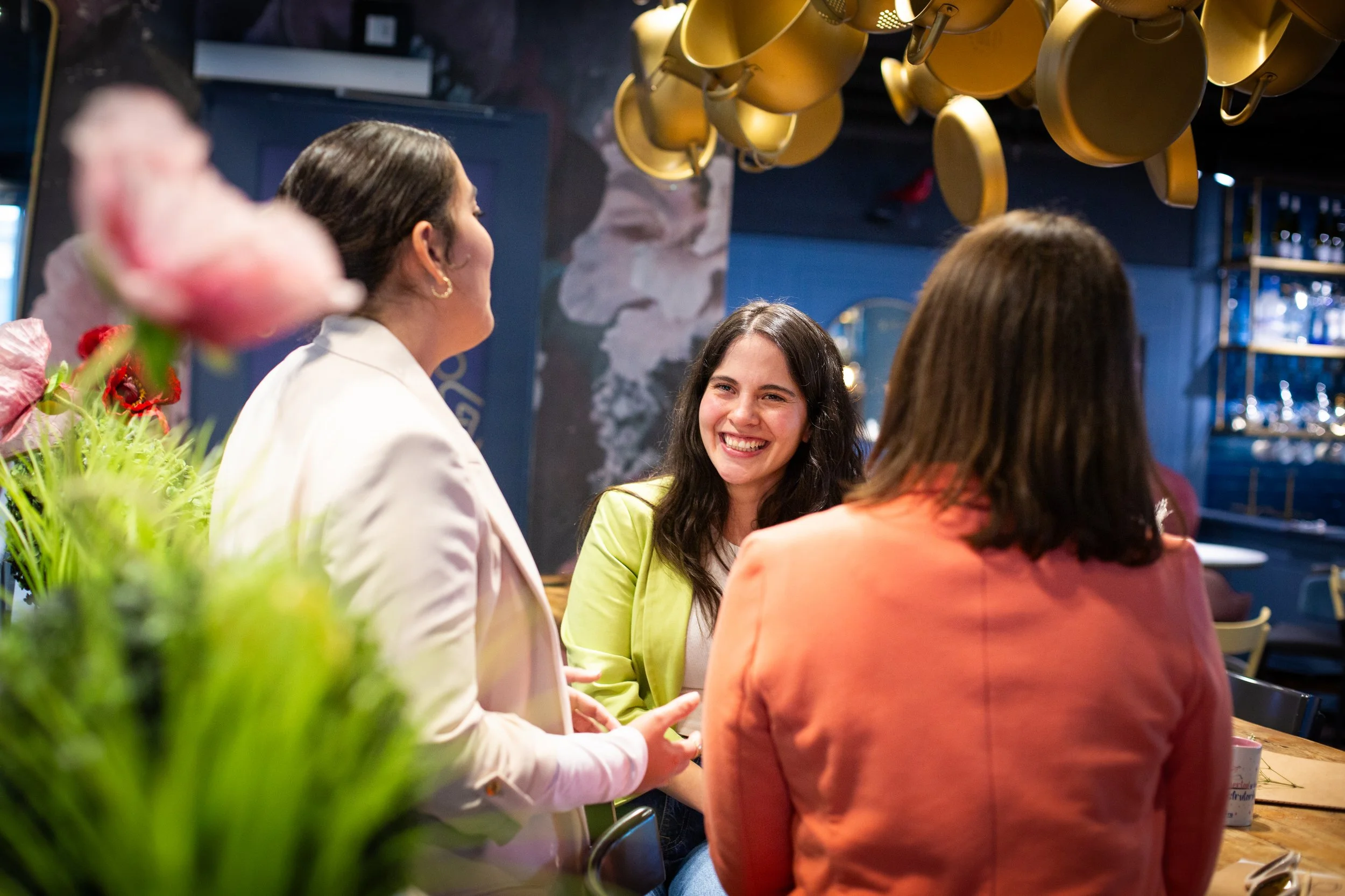 Three women engaged in conversation at a restaurant or cafe, with one woman smiling broadly. The background features hanging coffee cups and a bar area.
