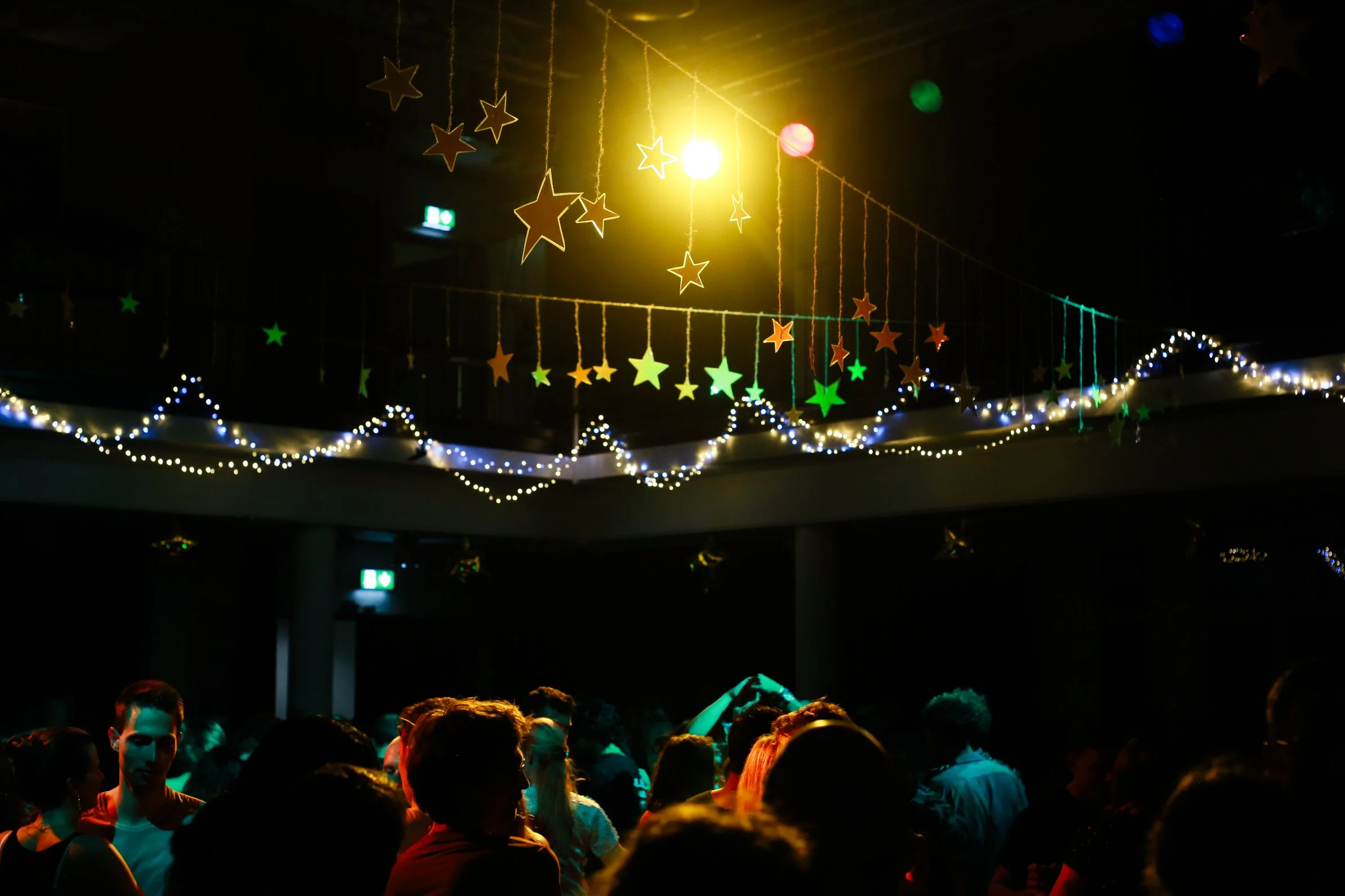 Dark indoor party or dance event with people, decorated with hanging yellow, orange, and green star decorations and string lights.