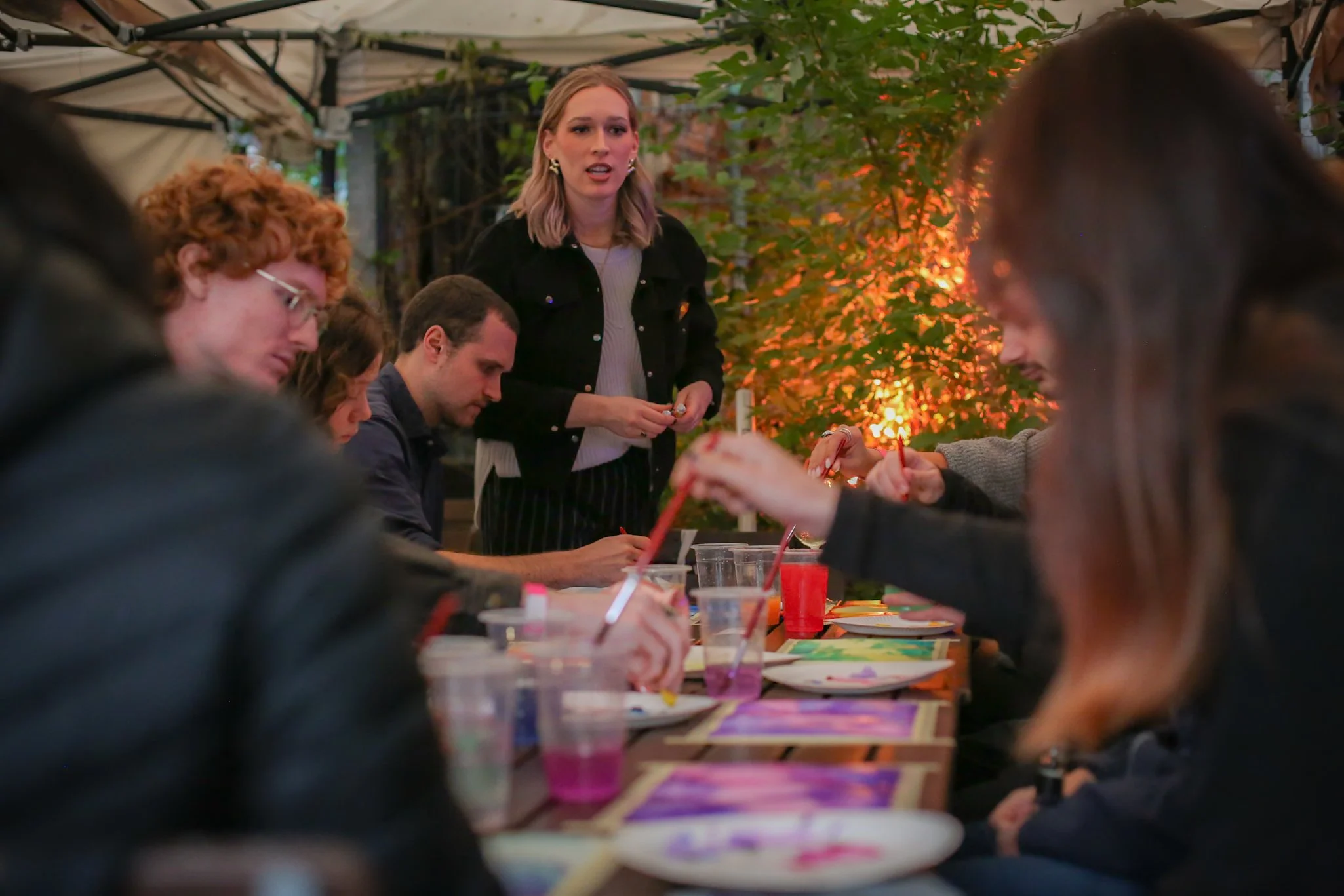 A group of people sitting at a long table outdoors at night, painting and decorating picture frames, with a woman standing and speaking to the group, under a canopy with string lights and surrounded by trees with orange leaves.