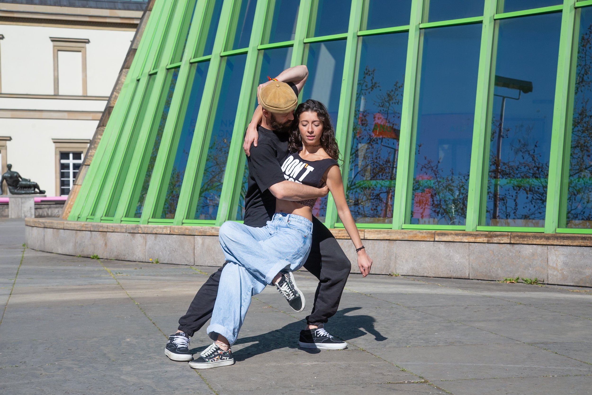 Two people dancing outdoors in front of a modern building with green window frames, on a paved surface.