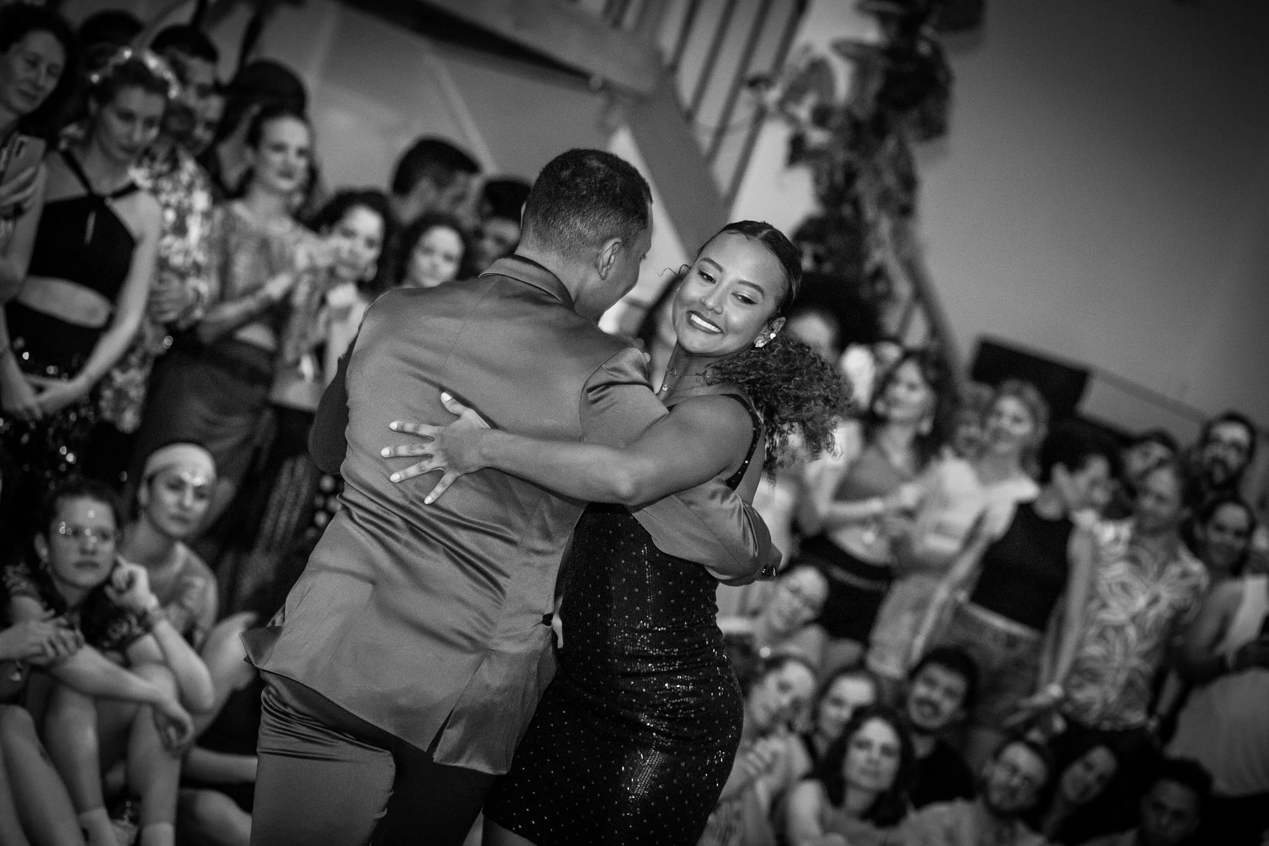 A couple dancing closely, with the woman smiling, at a crowded indoor event with people watching.