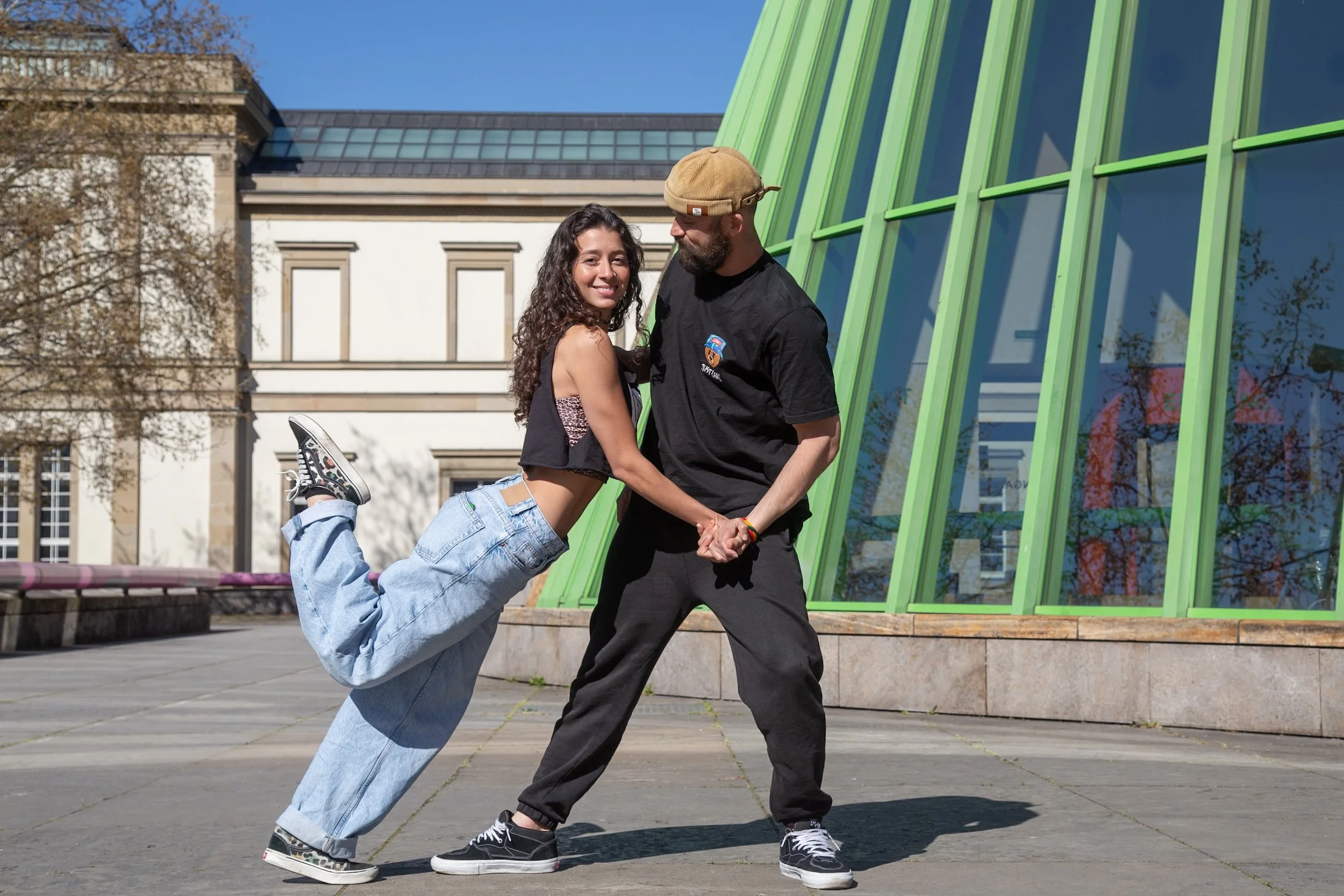 A young woman dancing with a man outdoors in front of a modern building with green window frames on a sunny day.