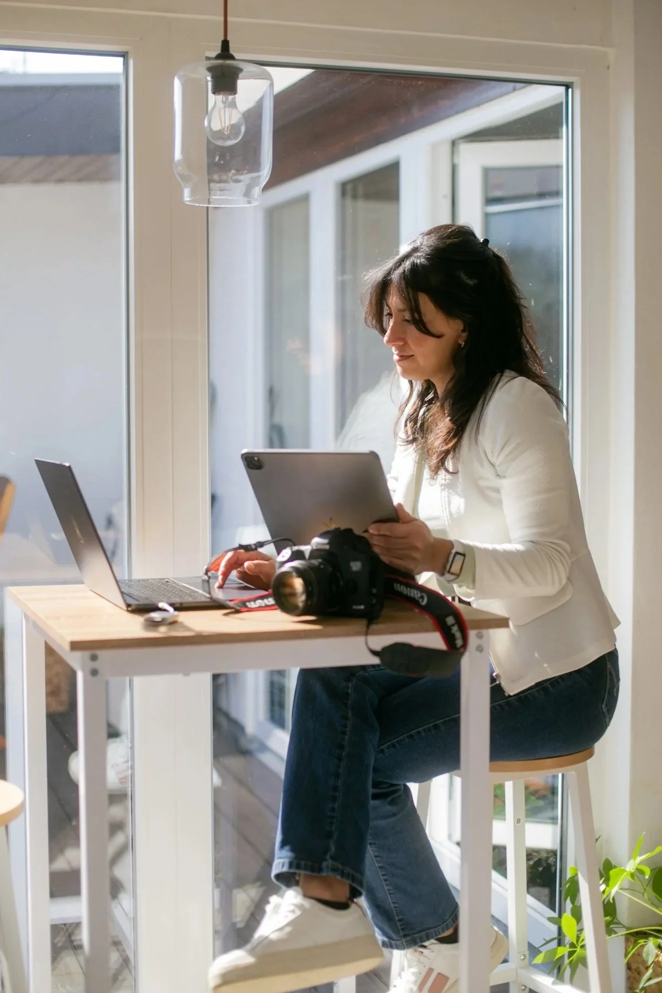 A woman sitting at a wooden table near a large window, working with a laptop, a camera, and a tablet.