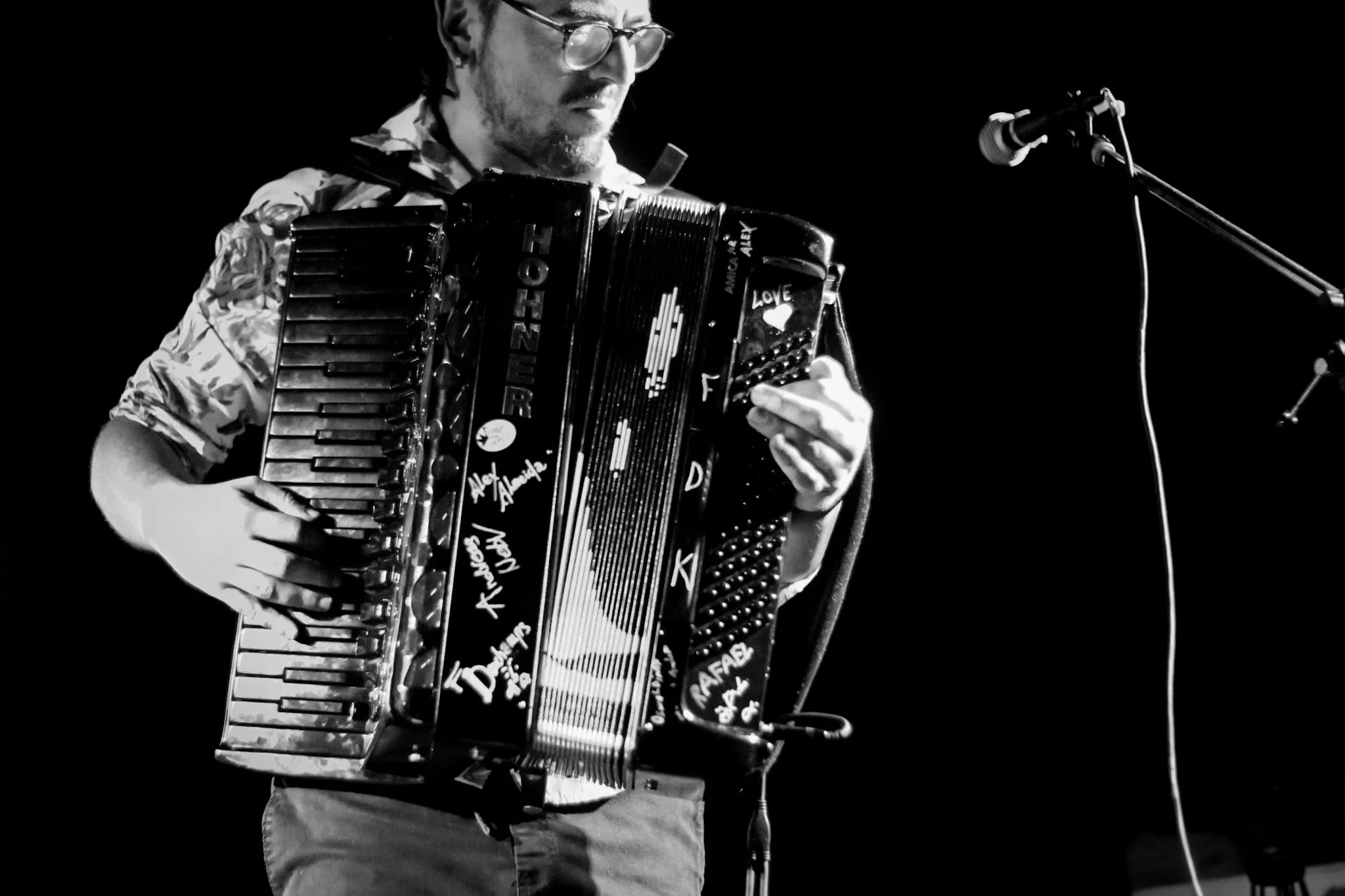 A man wearing glasses and a patterned shirt playing an accordion on stage, with a microphone nearby.