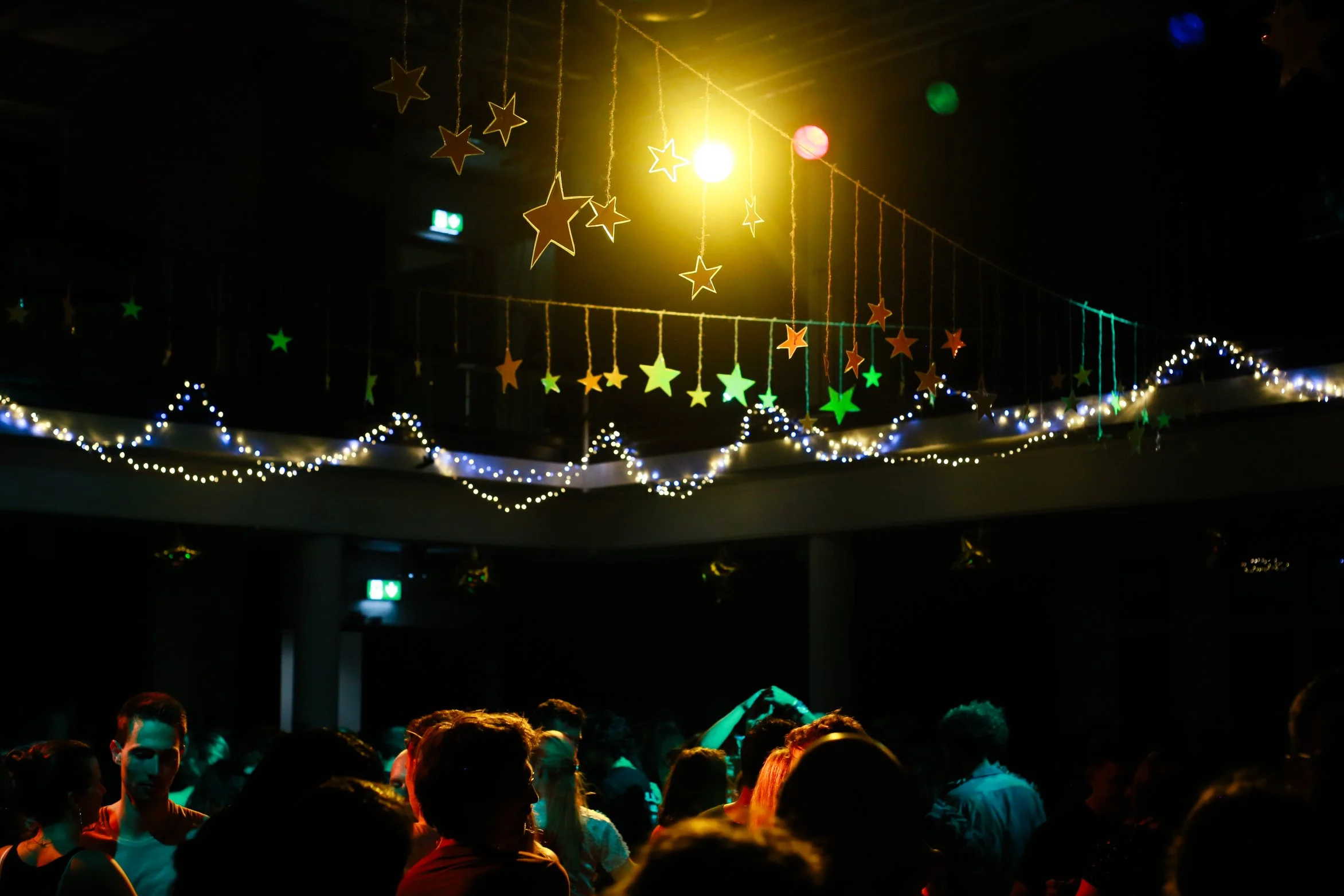 A crowd of people dancing or socializing at a party or event with colorful star-shaped decorations and string lights hanging from the ceiling, illuminated by a bright yellowish light.
