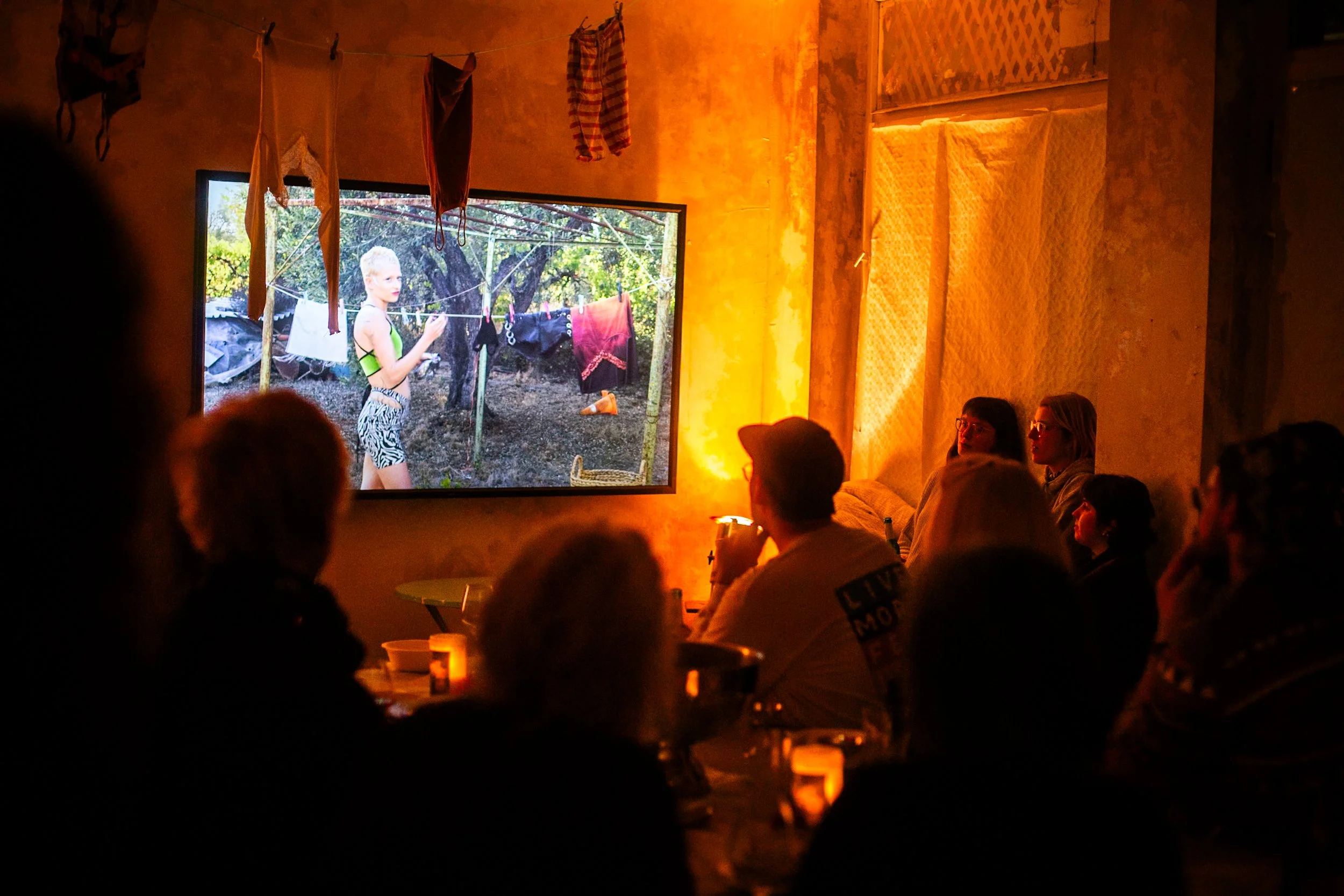 People watching a movie in a dimly lit room with candles on the table, while a large screen displays a woman outdoors hanging laundry.