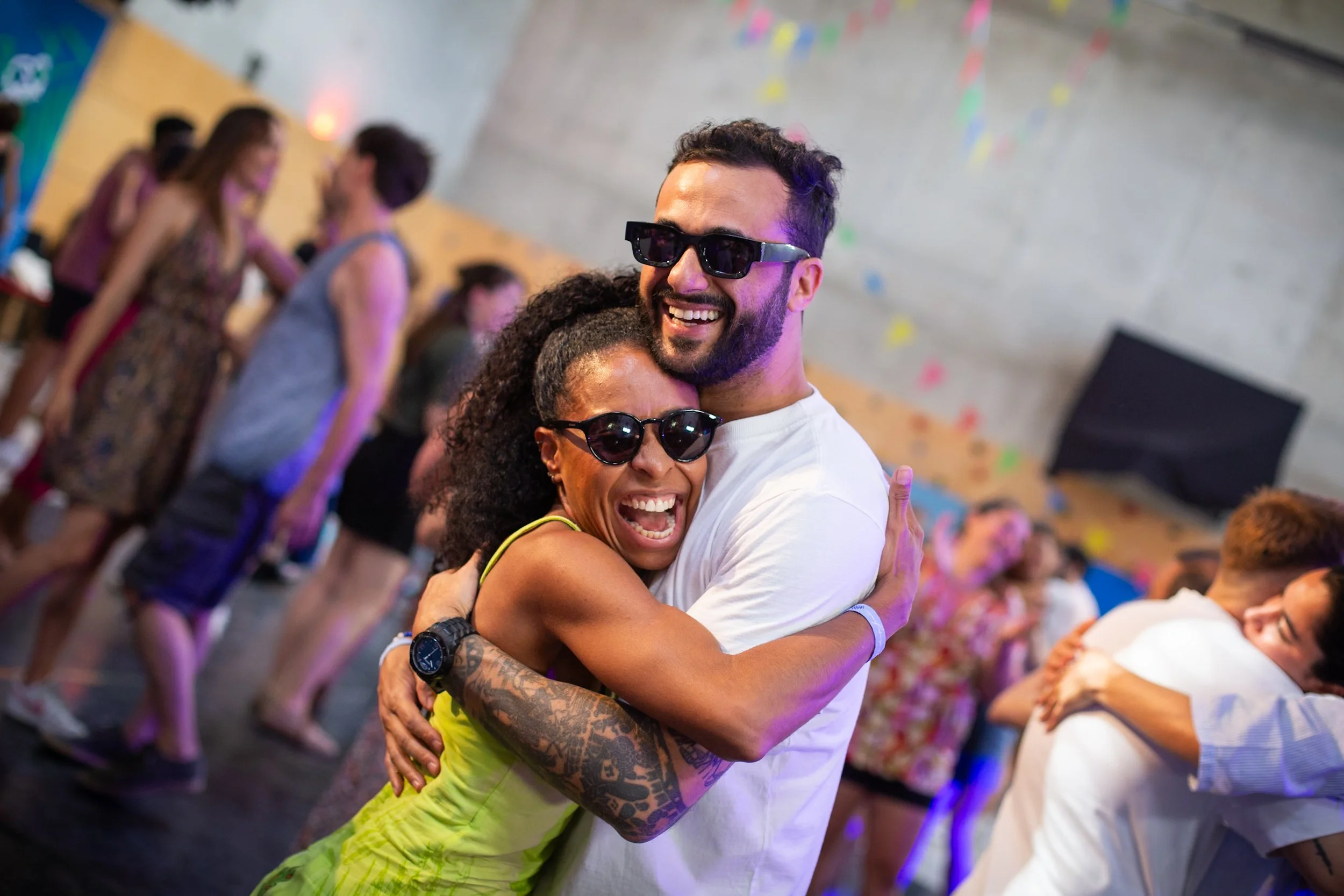 Two people wearing sunglasses hugging and smiling at a lively indoor dance party with colorful decorations and other dancing couples in the background.