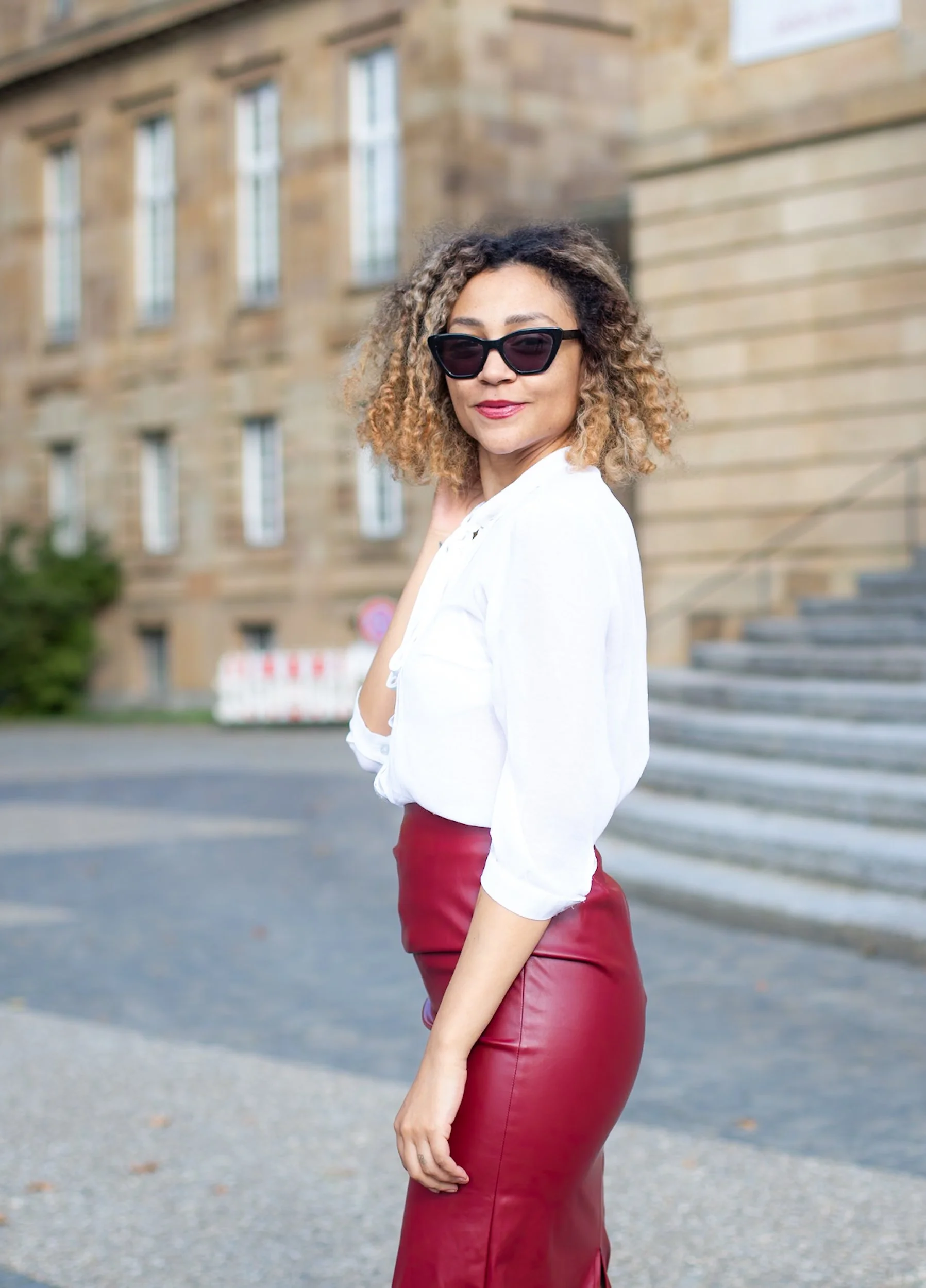 Stylish woman standing outdoors near stairs and a brick building, wearing sunglasses, a white blouse, and a red leather skirt.