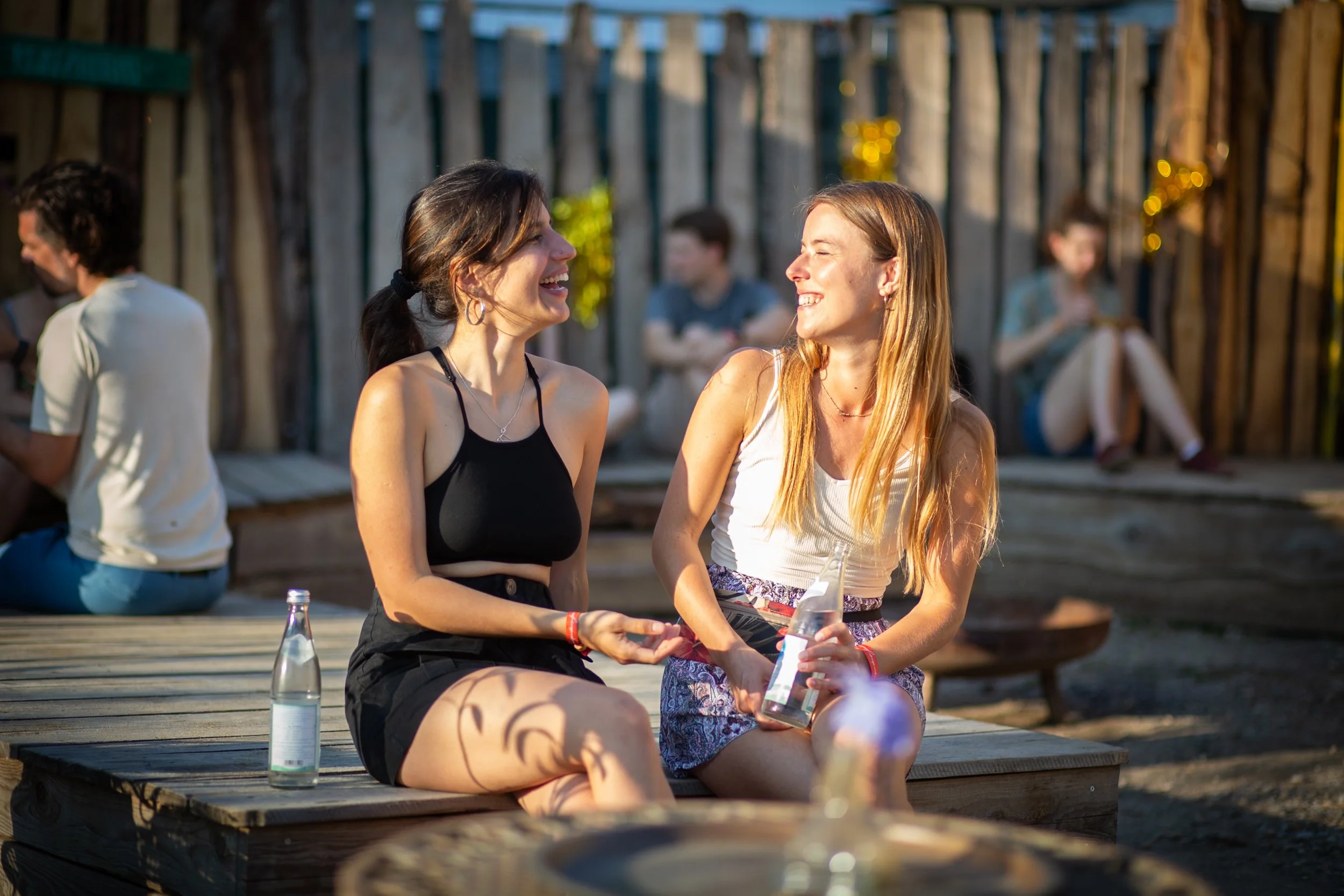 Two women sitting outdoors on a wooden platform, smiling and talking, with a small bottle of soda in one woman's hand. In the background, other people are also socializing in a rustic setting with wooden fencing and string lights.