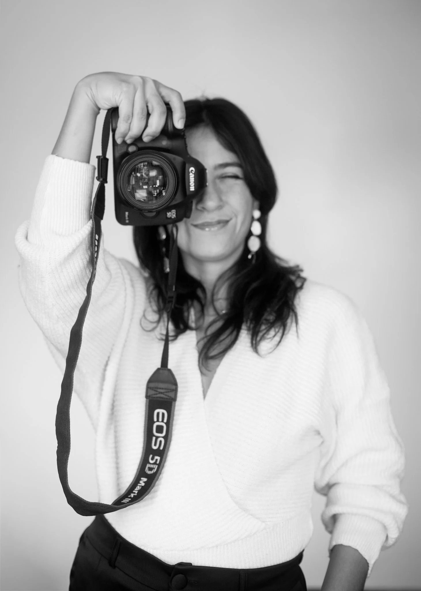 Black and white photo of a woman with dark hair, smiling, holding a Canon camera in front of her face. She is wearing a light-colored sweater and earrings.