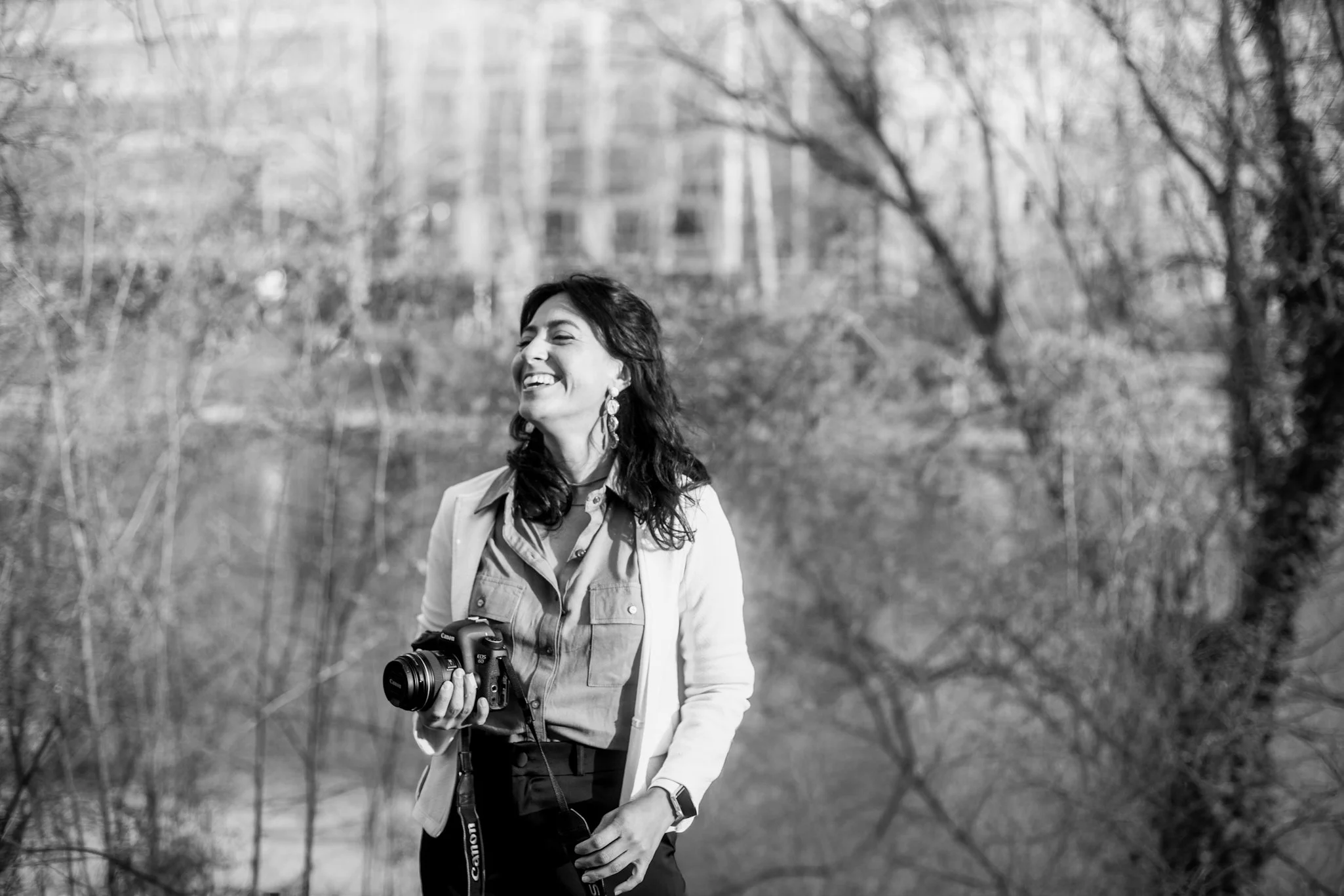 A woman with dark hair, smiling and holding a camera, standing outdoors in a park with trees in the background.