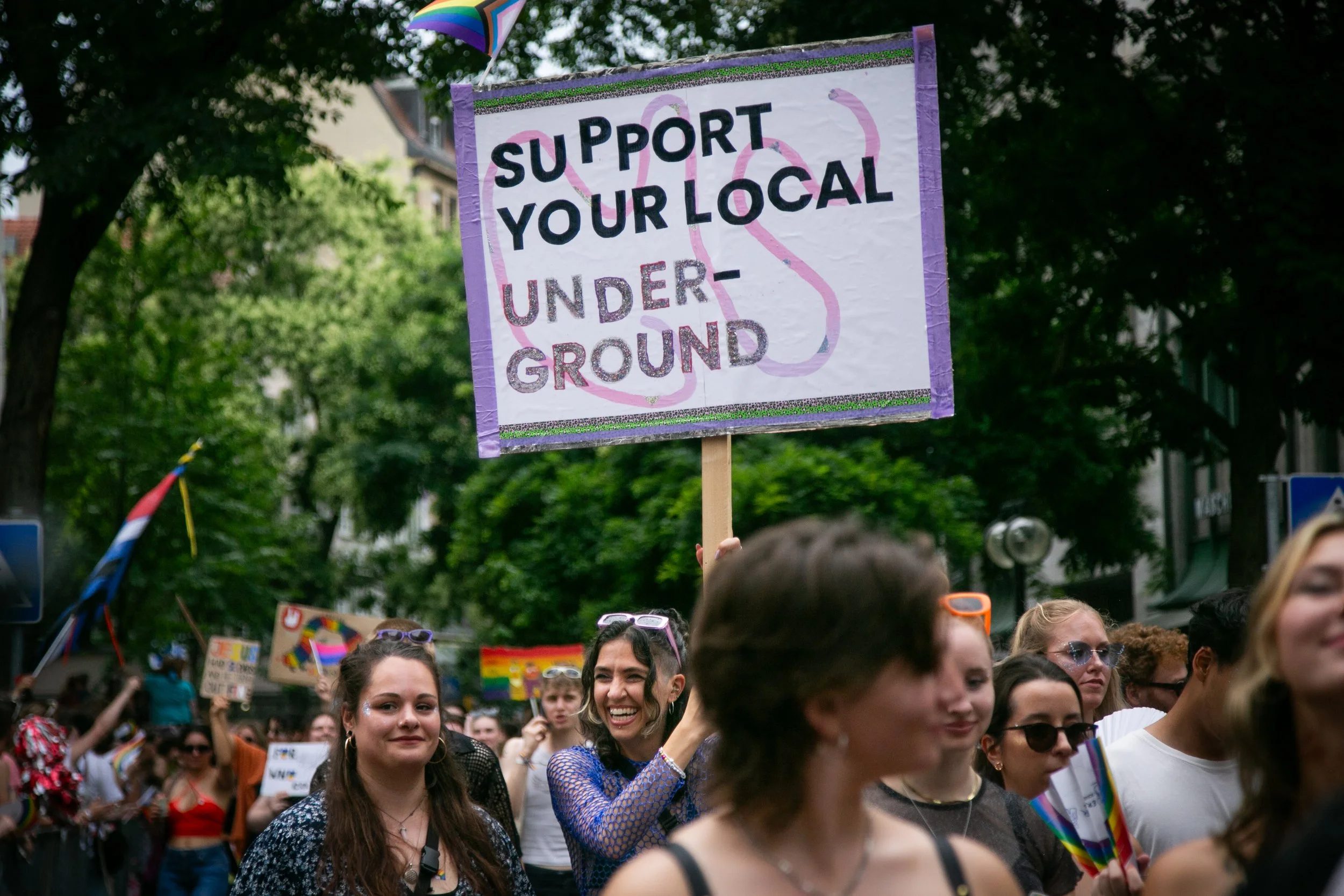 People participating in a pride parade or rally, holding signs and rainbow flags. One woman in the center is smiling and holding a sign that says 'Support Your Local Underground' with rainbow and glitter decorations.