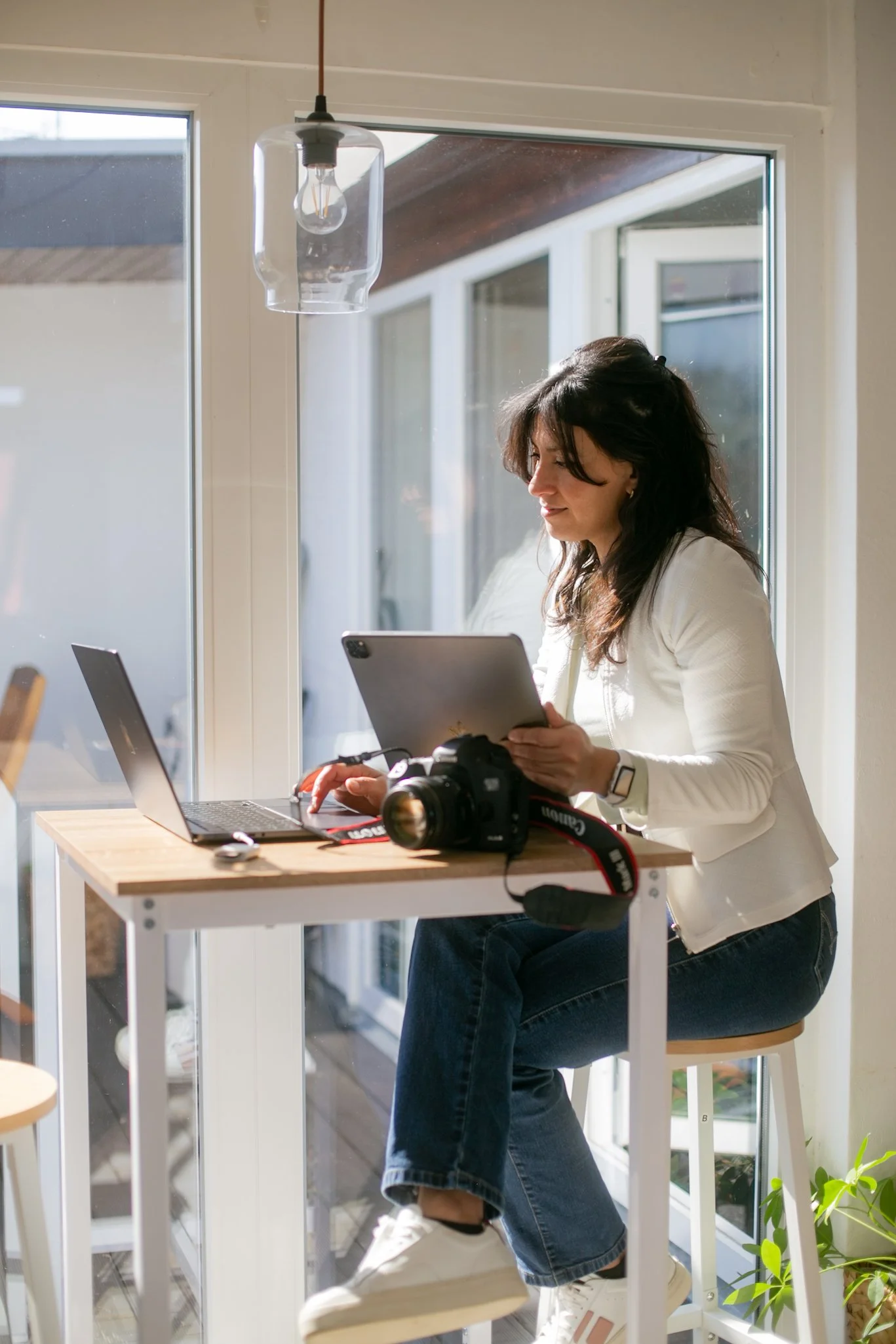 Woman sitting at a small wooden desk near a window, working on a tablet. There is a laptop, a professional camera, and a camera strap on the desk. The room is bright with natural sunlight.