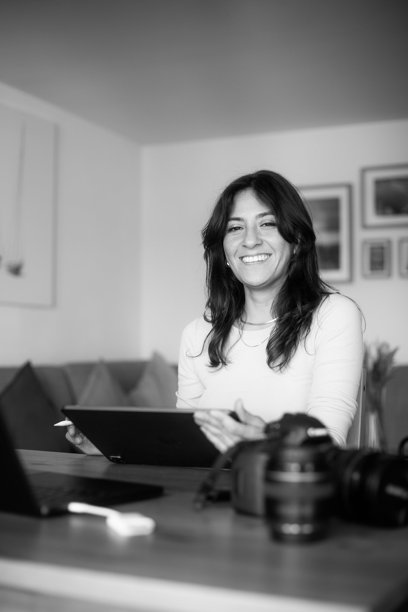 A woman smiling while sitting at a desk with a laptop and camera, in a room with framed pictures on the wall.