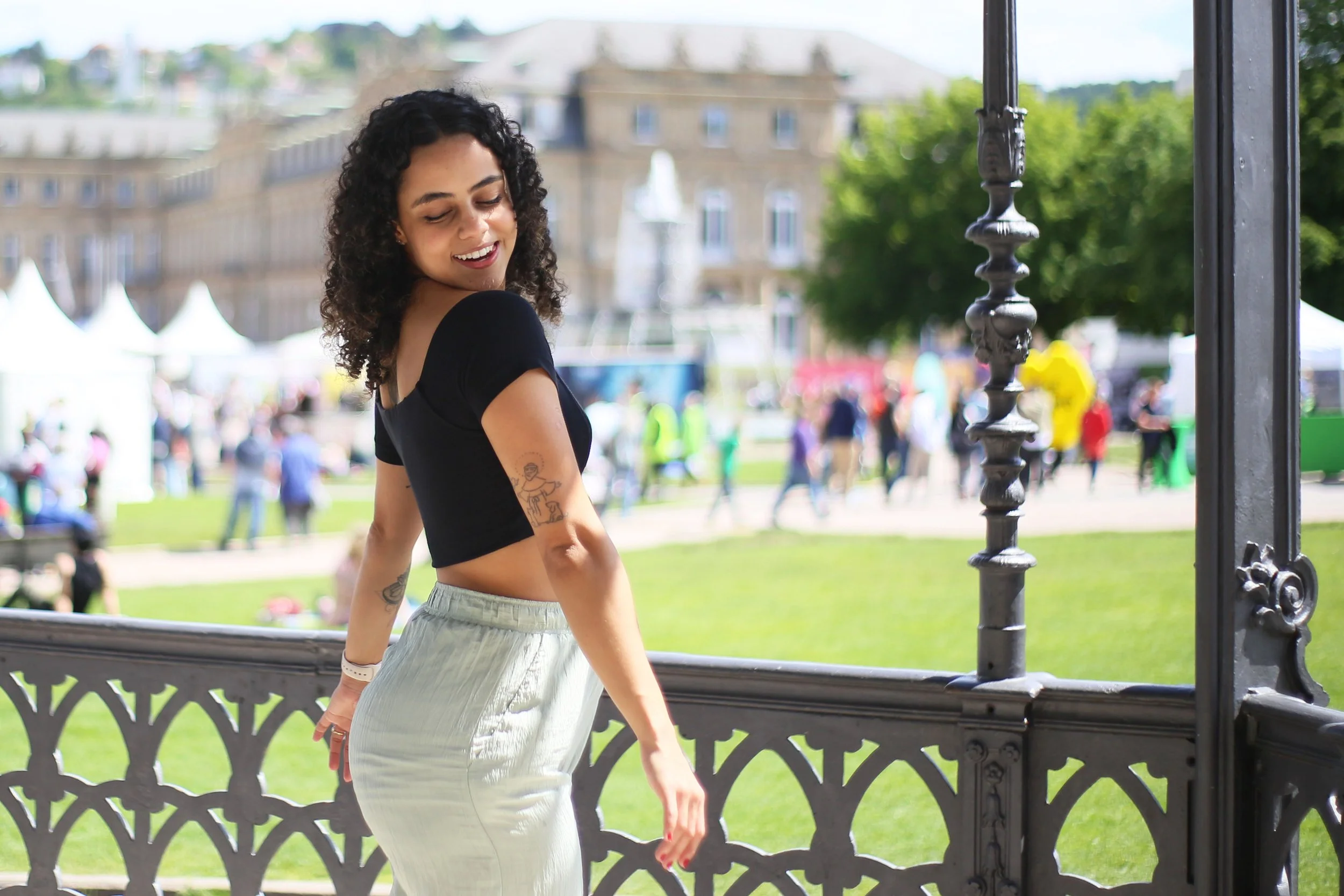 A woman standing on a decorative metal bridge in a park, smiling and looking down, with a crowded park and historic buildings in the background on a sunny day.