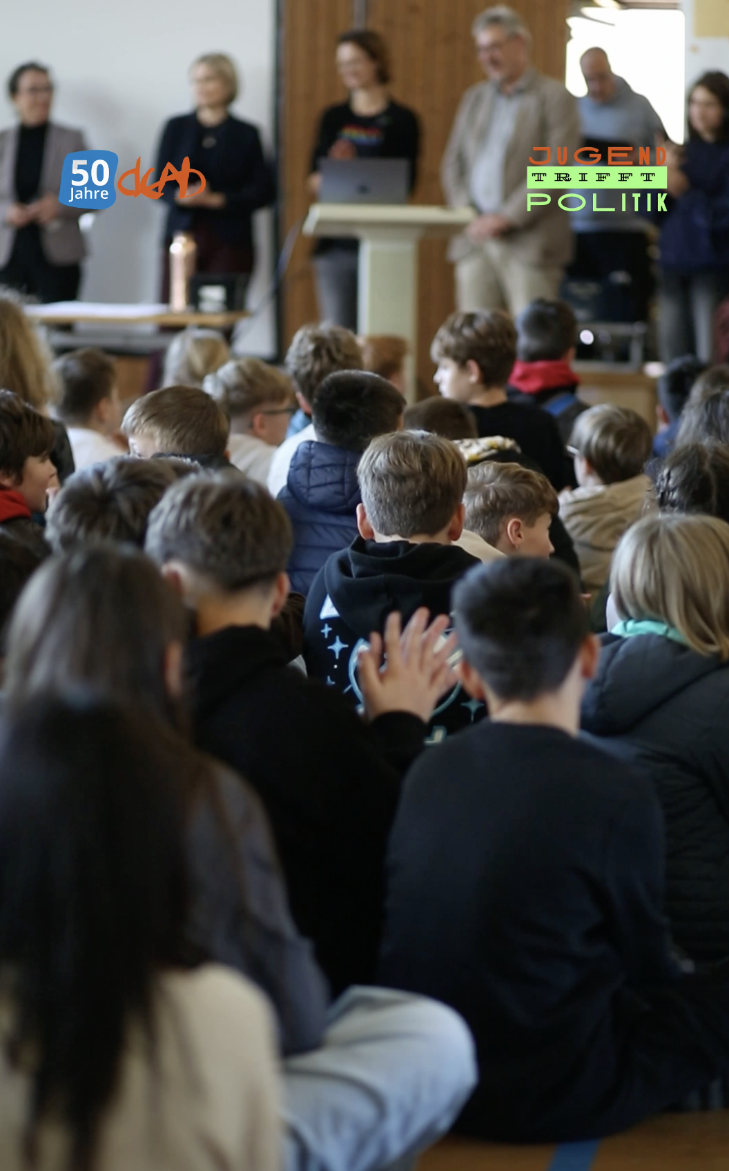 Group of young students attending an indoor event with several adults standing at a podium or front of the room, some of whom are speaking or presenting.