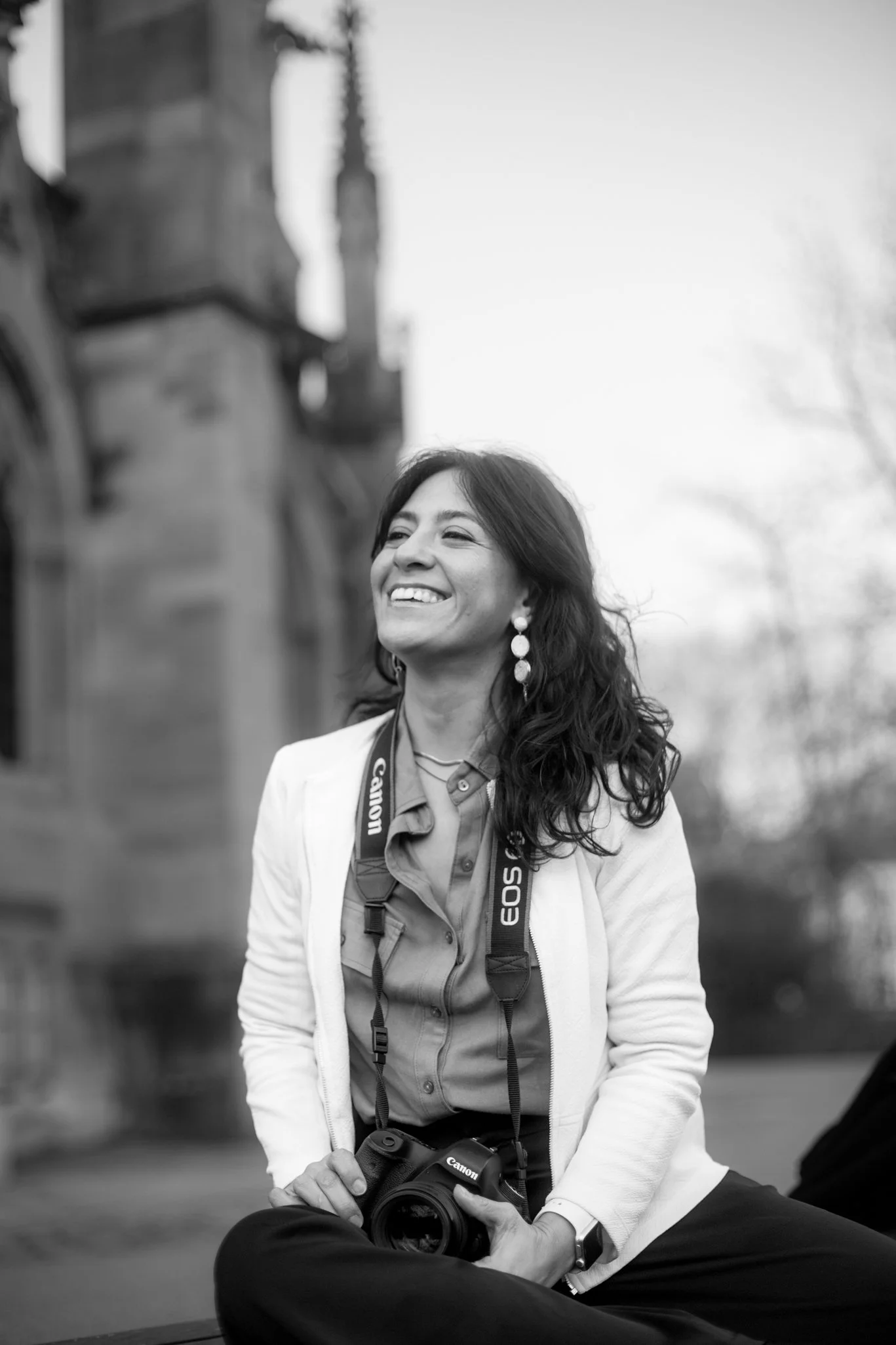 A woman sitting outdoors smiling, holding a Canon camera with a strap hanging around her neck, wearing earrings, a shirt, and a blazer, with an old building in the background.