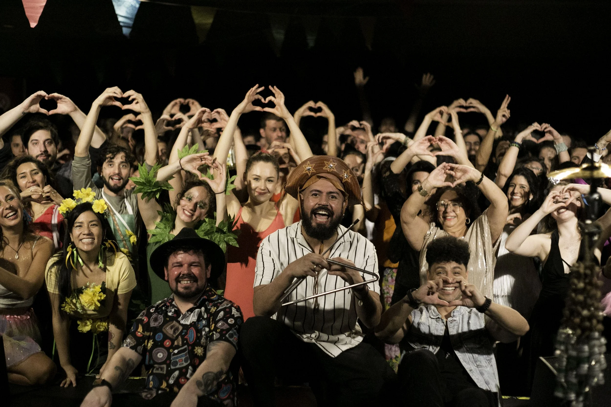 A diverse group of people at a celebration, making heart shapes with their hands, some dressed in costumes and accessories, smiling and enjoying the event.