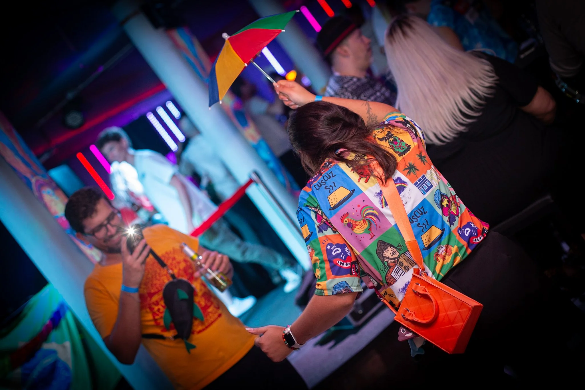 People at a colorful neon-lit party, with one person holding a rainbow umbrella and others taking photos.