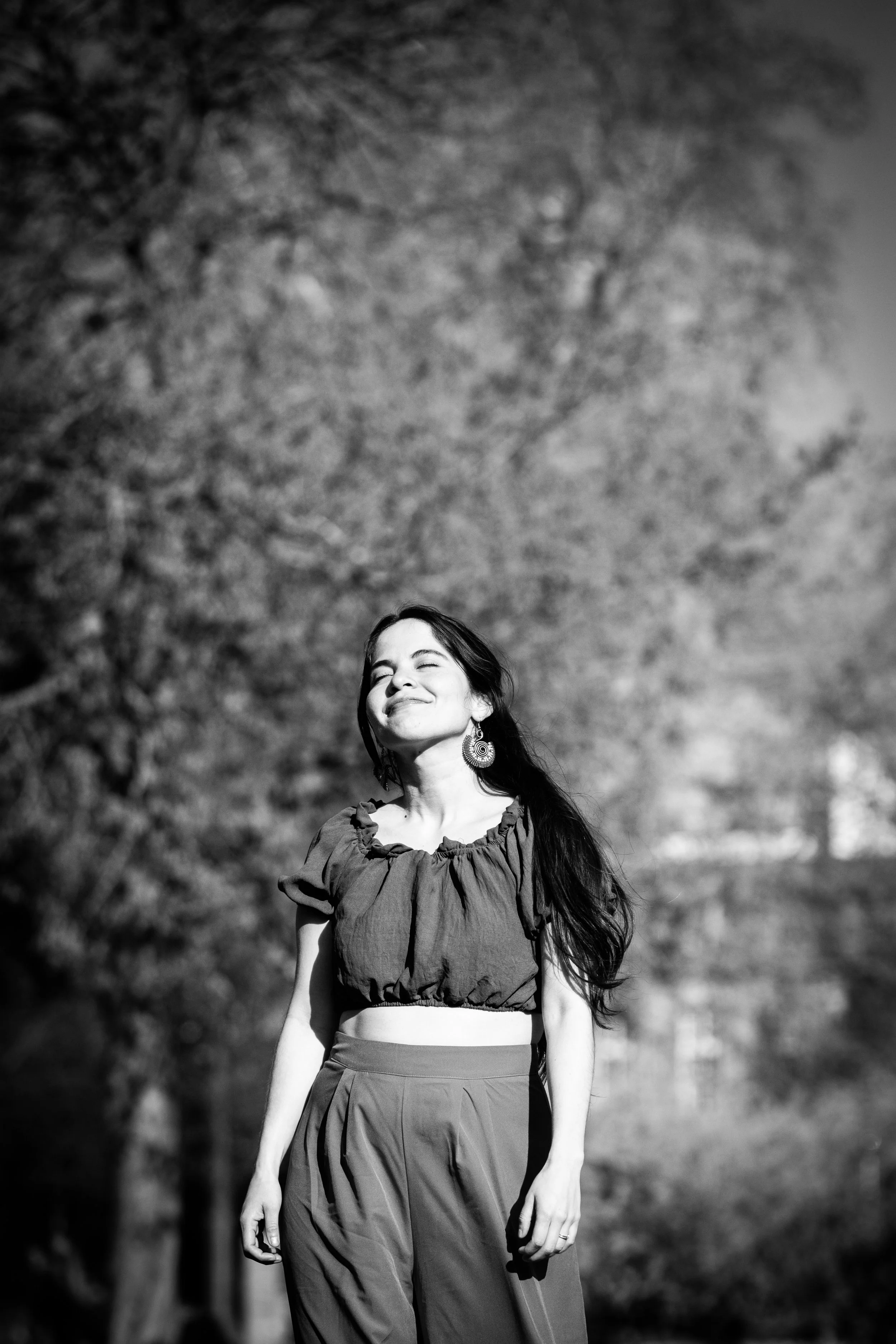 A woman is enjoying the sunshine outdoors, wearing a cropped top and high-waisted pants, with her eyes closed and a smile on her face.
