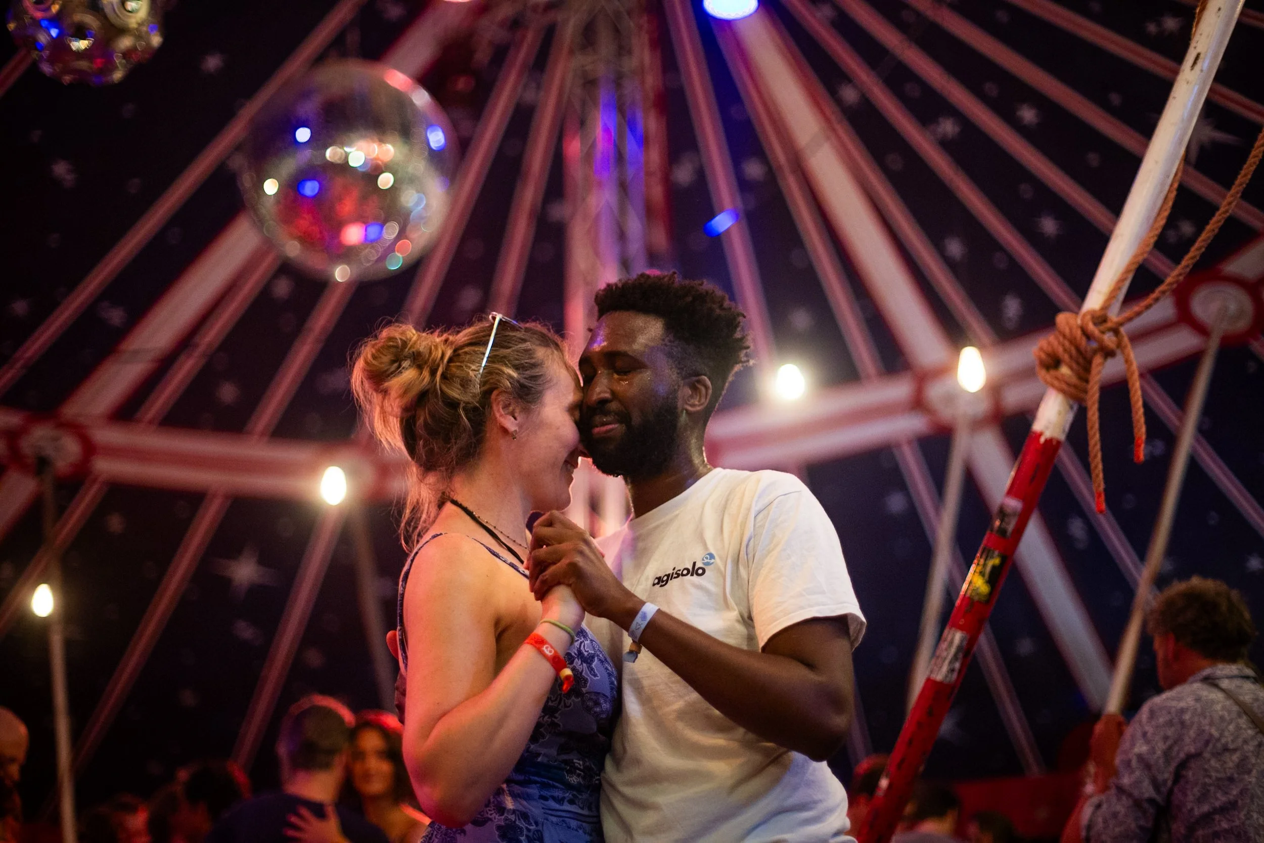 A couple is dancing closely together inside a carnival ride with a Ferris wheel in the background at night.